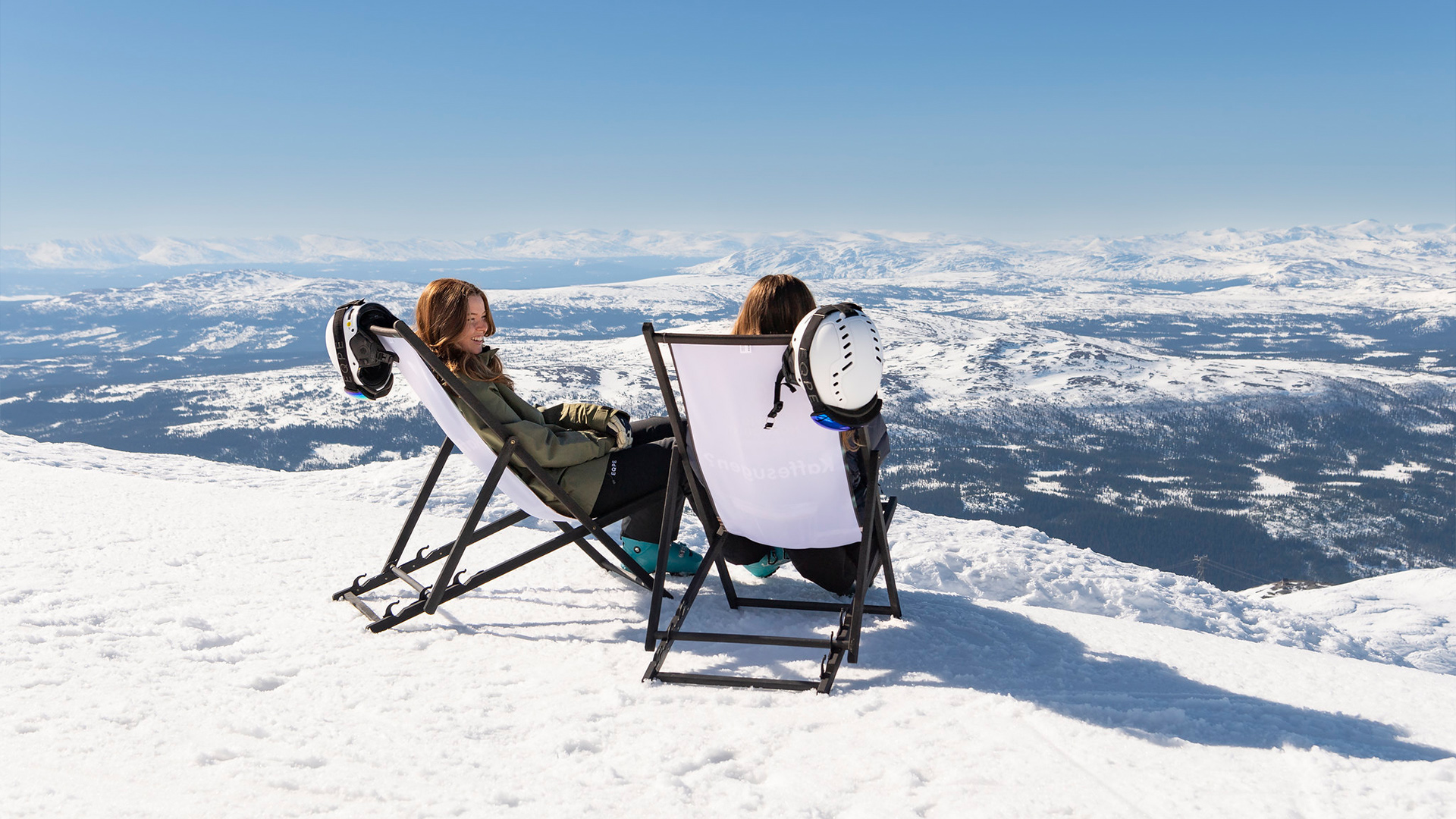 Två personer njuter av solen i solstolar på snötäckt bergstopp med panoramautsikt över fjällandskap i Åre, Sverige