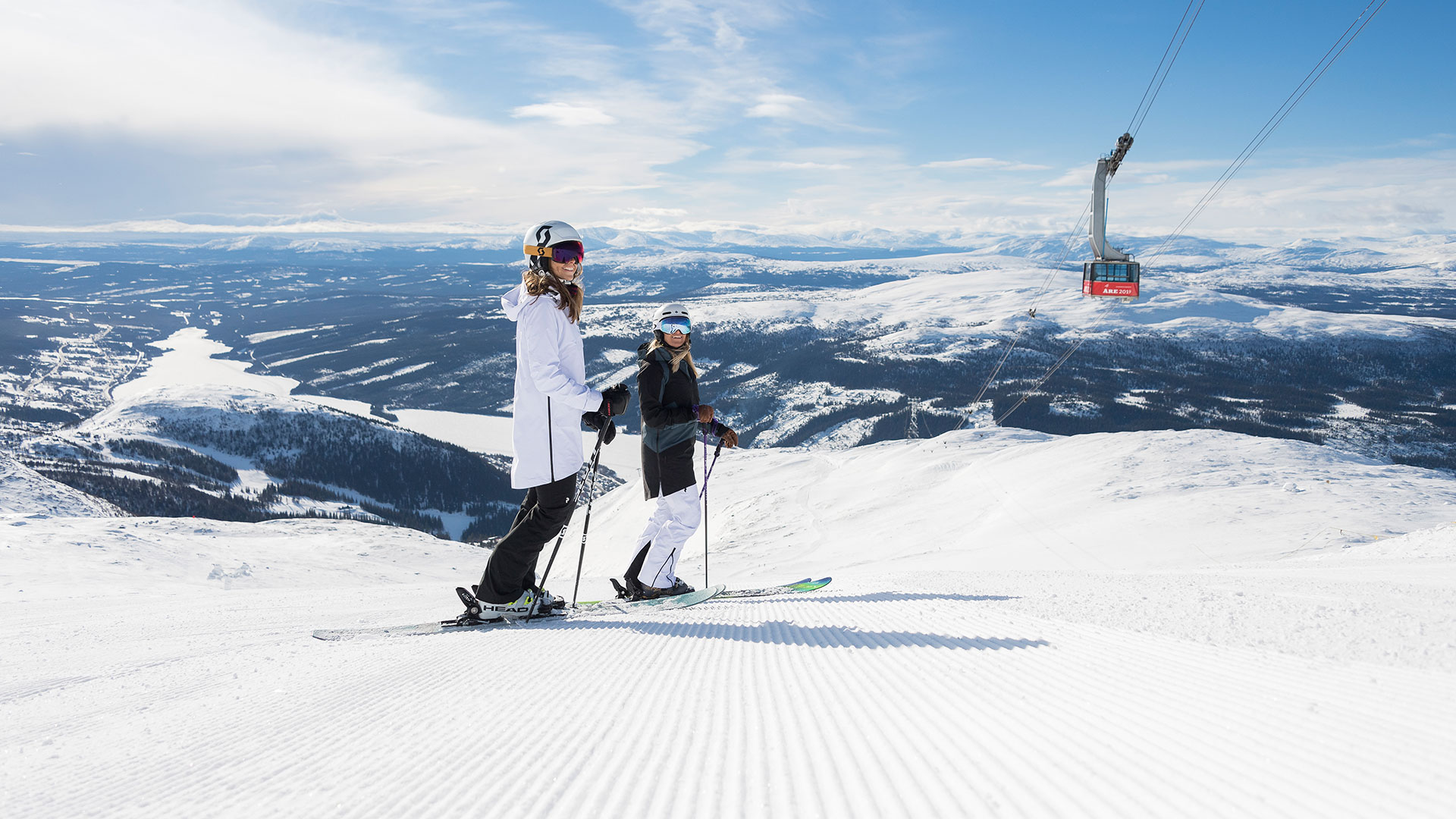 Två skidåkare i vinterlandskap i Åre, Sverige, med kabinbanan i bakgrunden. Snötäckta fjäll och klarblå himmel skapar en vacker vy