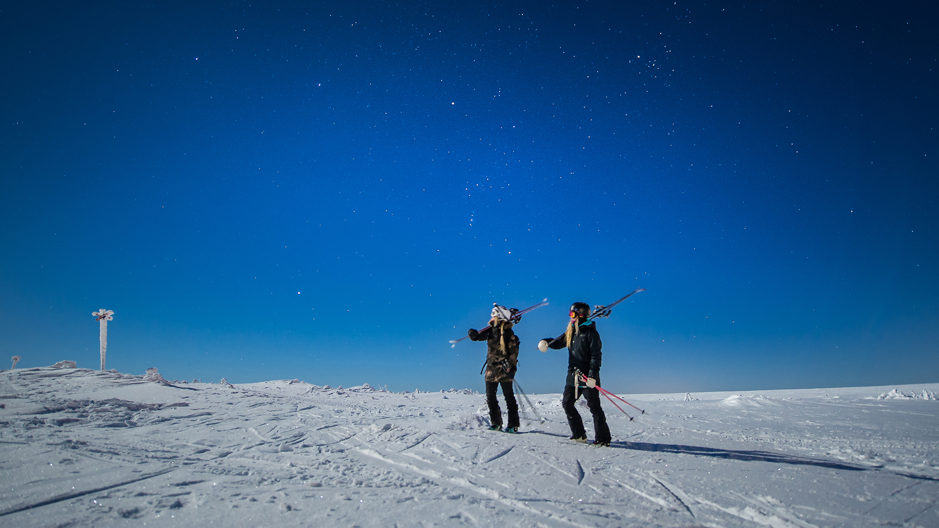 To personer med ski på skuldrene står på en snedækket højderyg under en klar nattehimmel med stjerner