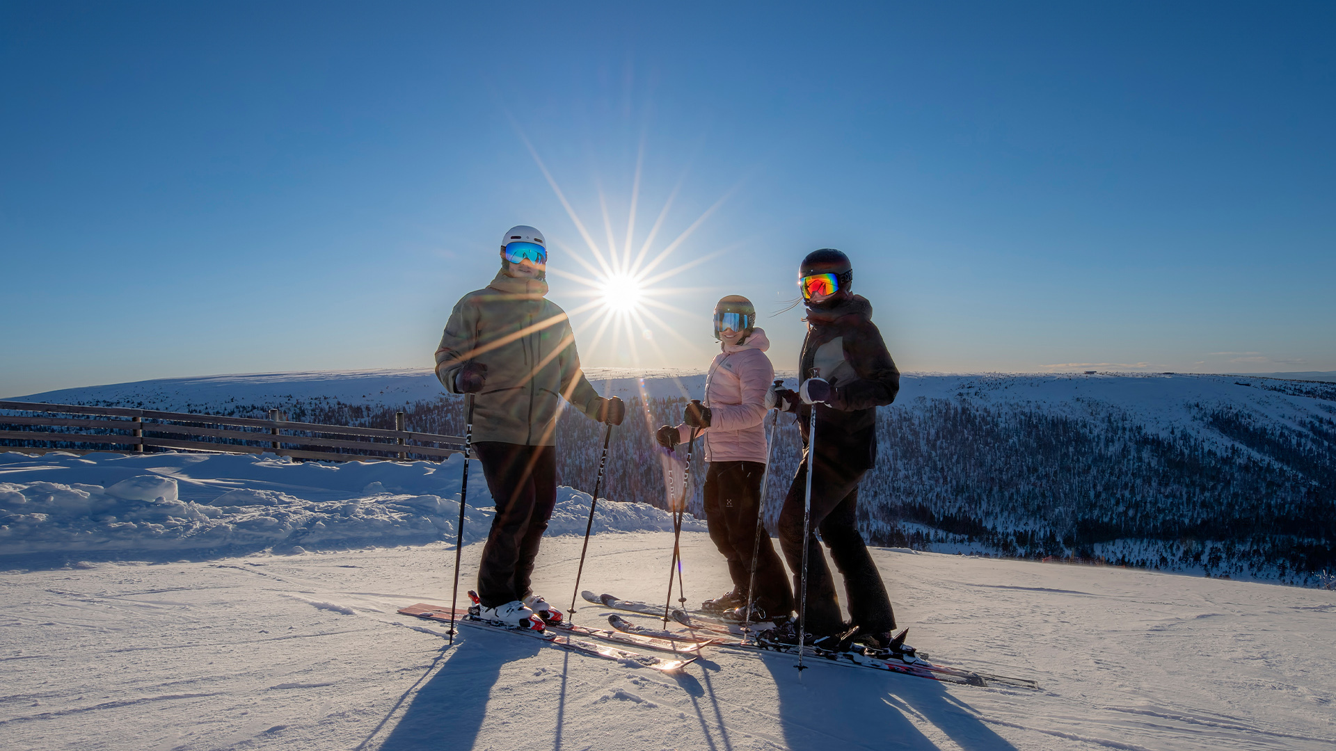 Tre skidåkare på en snötäckt bergstopp med solen i bakgrunden, klarblå himmel och vidsträckt utsikt över vinterlandskap