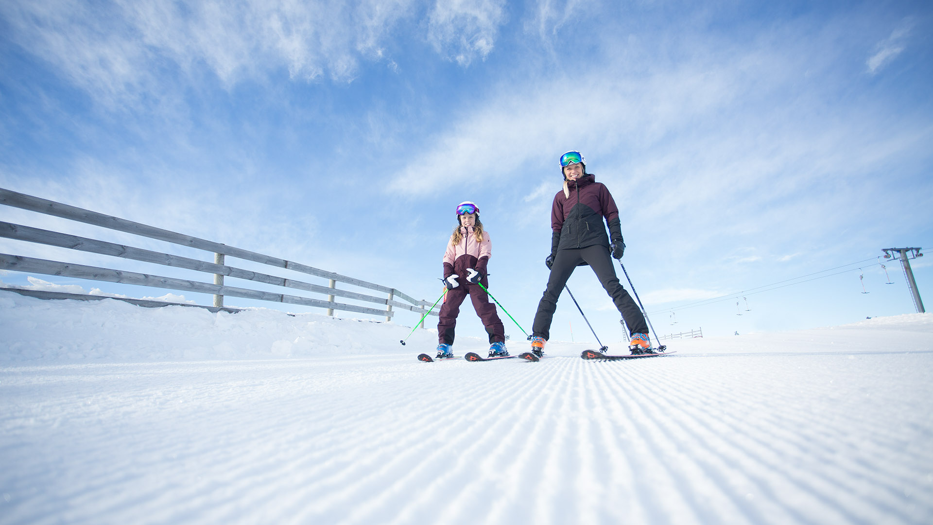Två personer åker skidor på en solig dag i en snötäckt skidbacke med blå himmel i bakgrunden