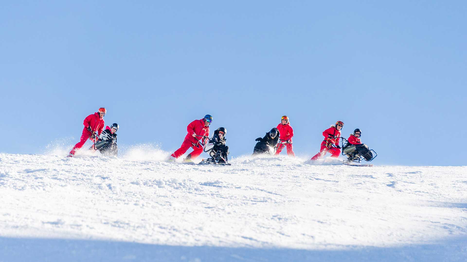 Skidinstruktörer i röda kläder assisterar personer på sit-skis nerför snöig backe under klarblå himmel