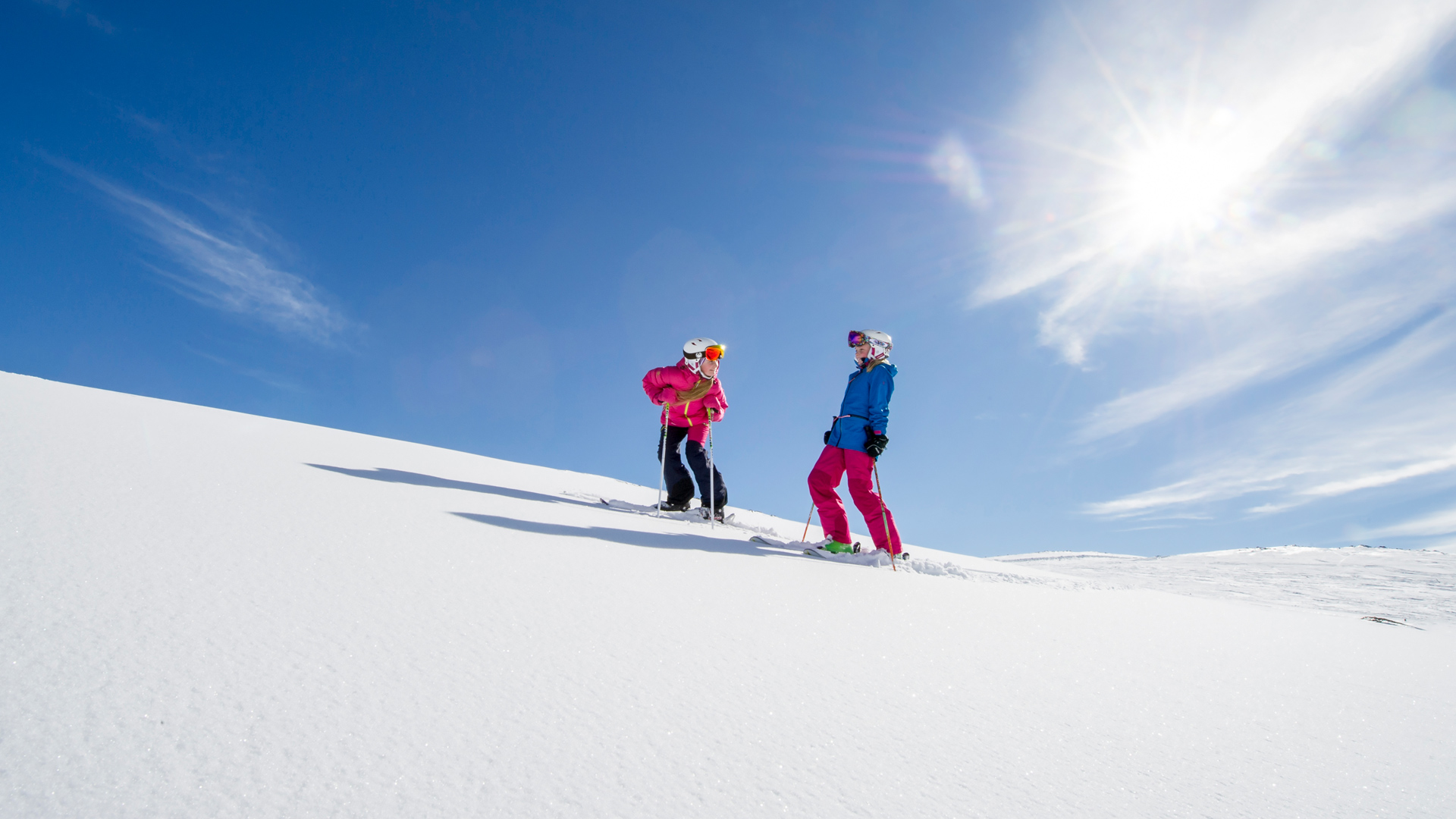 Två personer som åker skidor i en snötäckt backe under klarblå himmel med solsken
