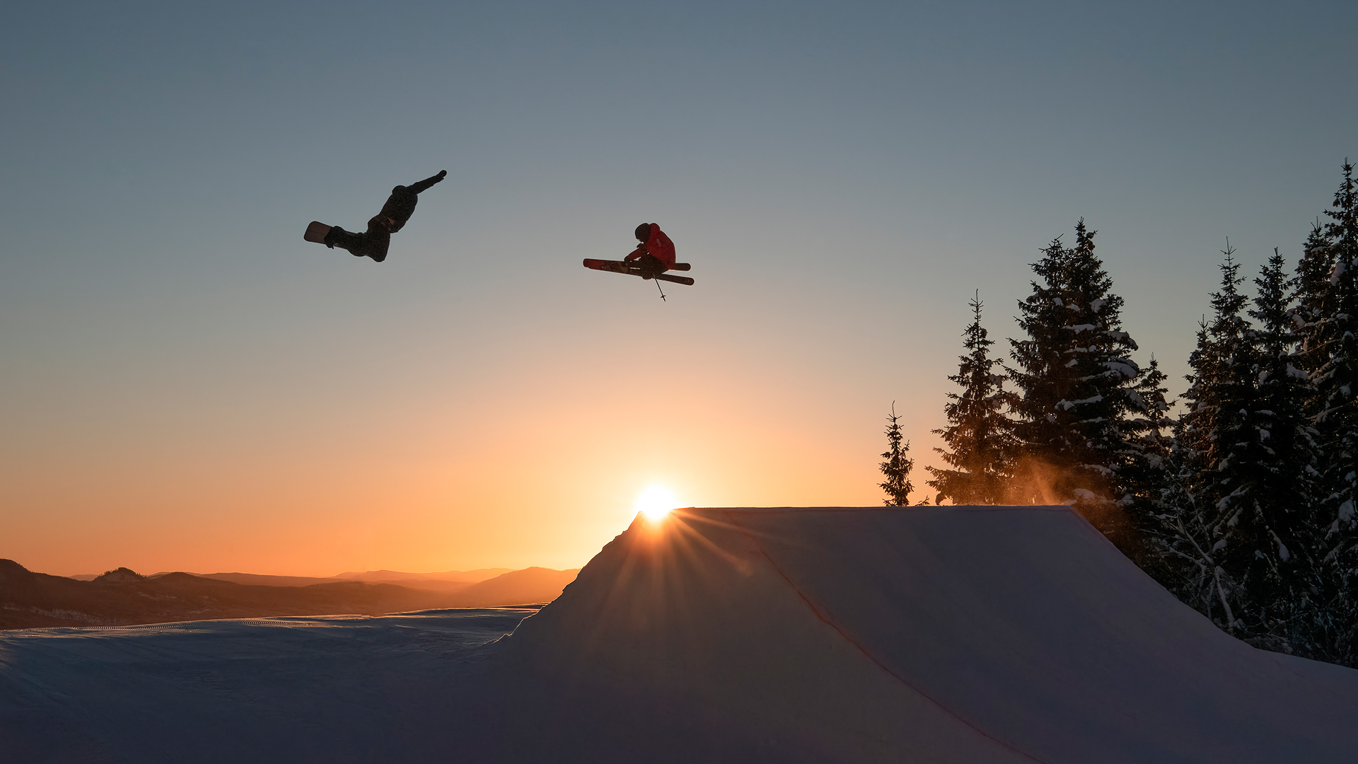 Snowboardåkare och skidåkare utför imponerande hopp i solnedgången, omgivet av snötäckta träd