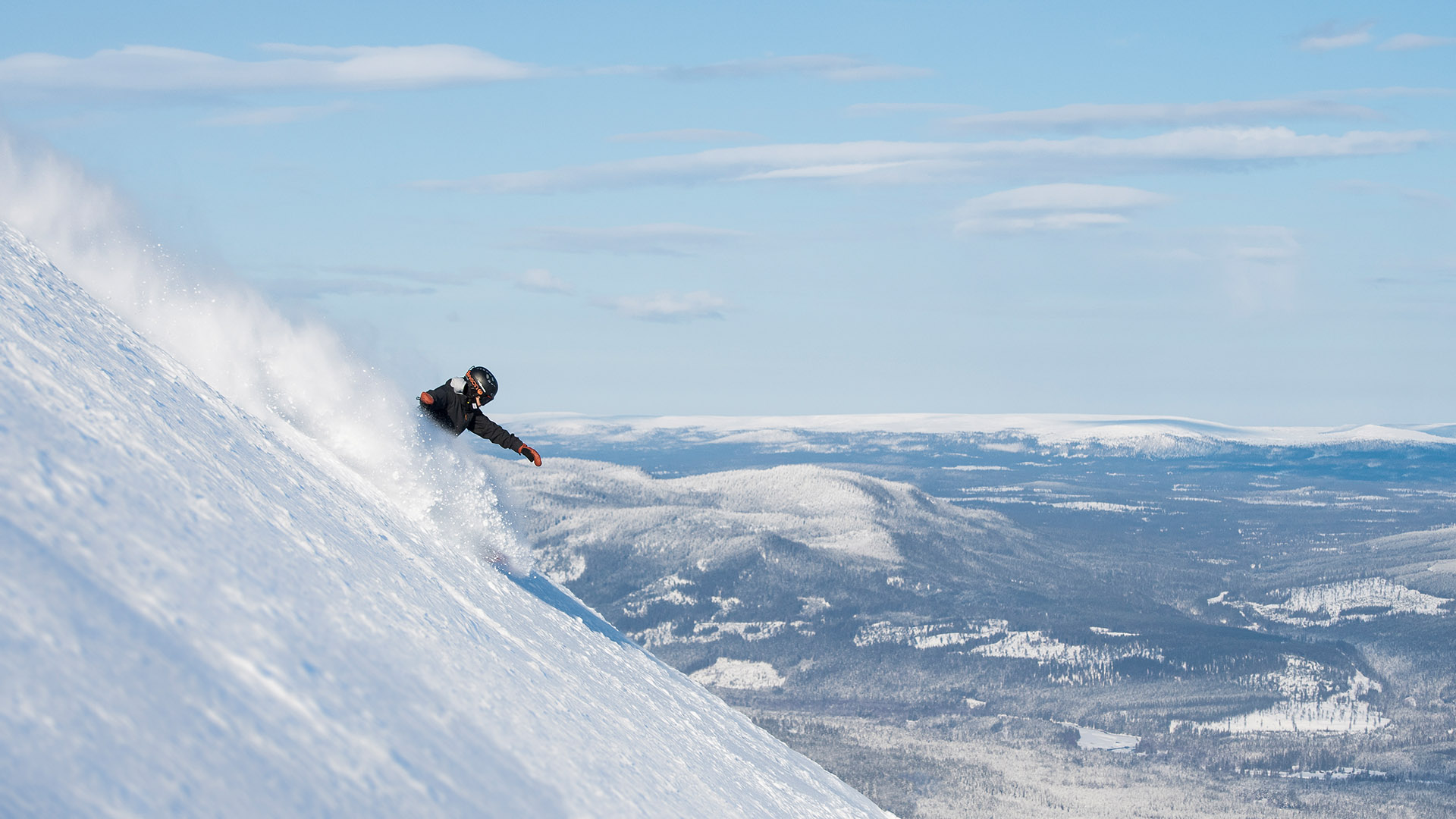 Snowboardkjørere suser nedover snødekket bakke med utsikt over fjellandskap i solen