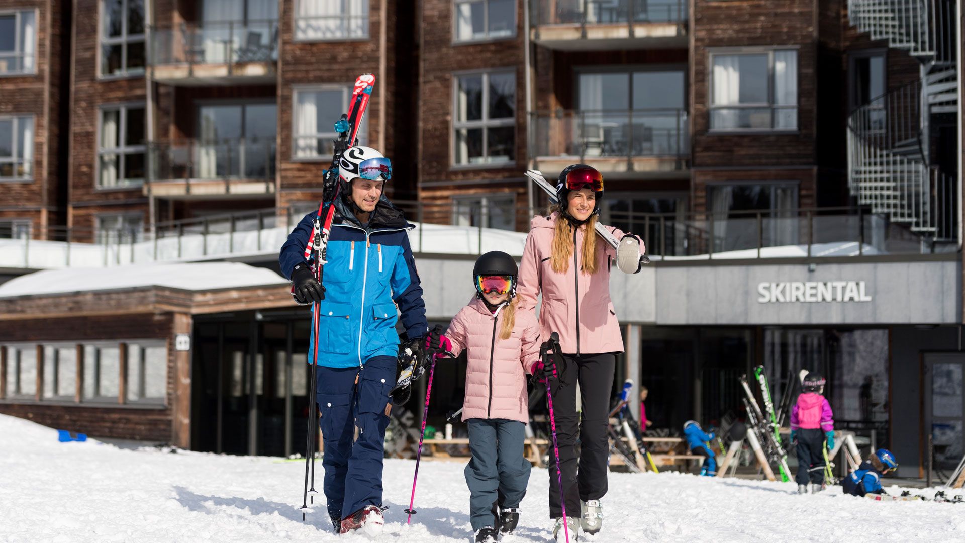 Familj på skidsemester bär skidutrustning utanför skidhyra i vinterlandskap