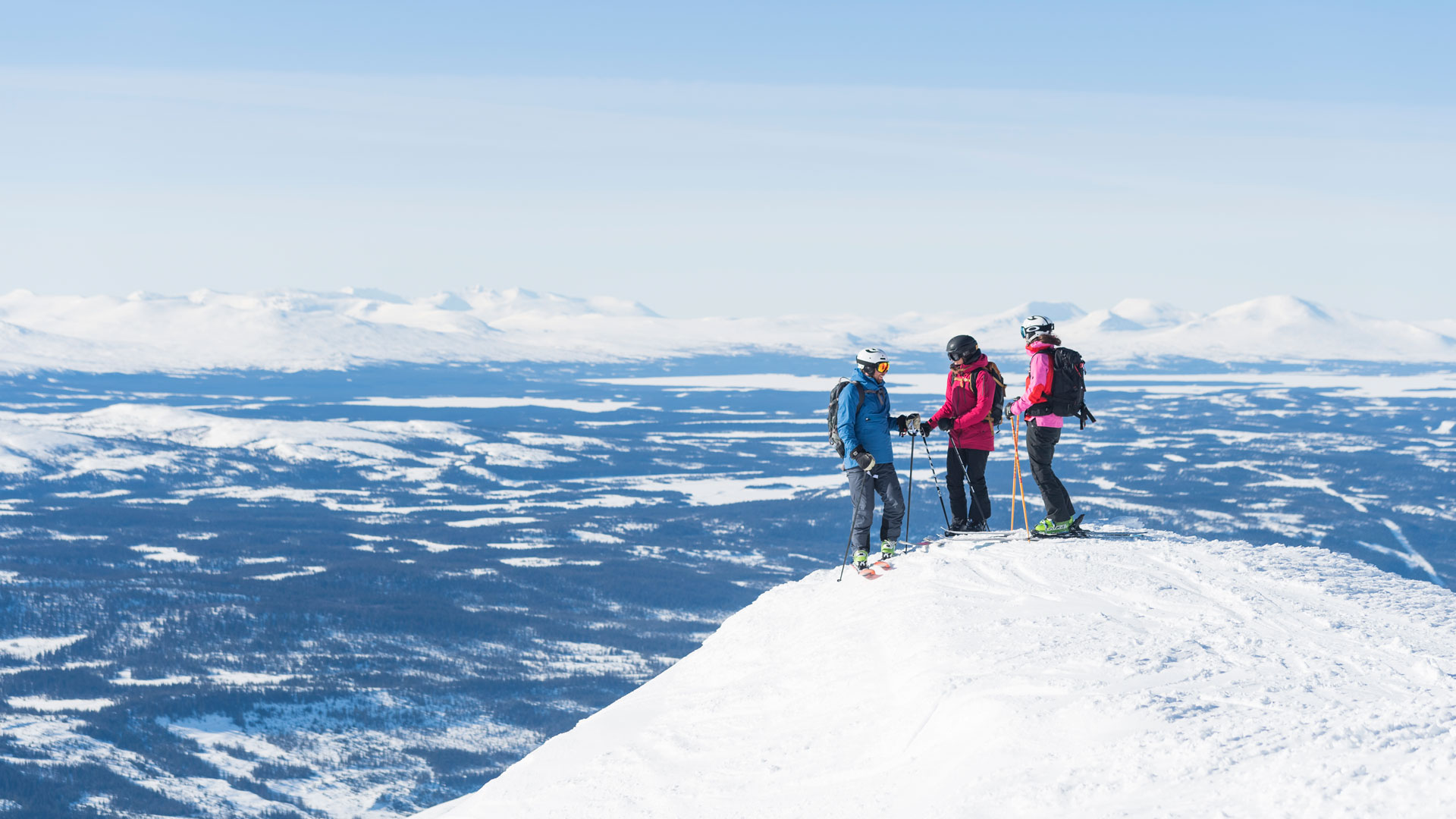 Tre personer, der står på ski på en snedækket bjergtop i fjeldene under en klarblå himmel
