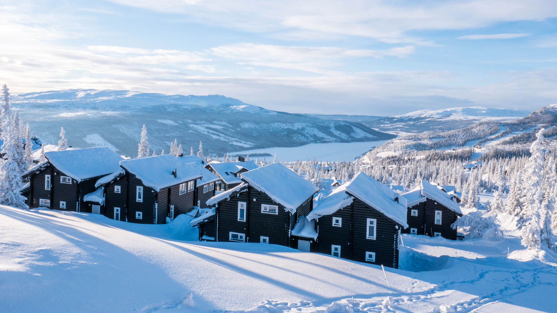 Snötäckta timmerstugor i bergslandskap med sjö och fjäll i bakgrunden under klar vinterdag i Åre, Sverige