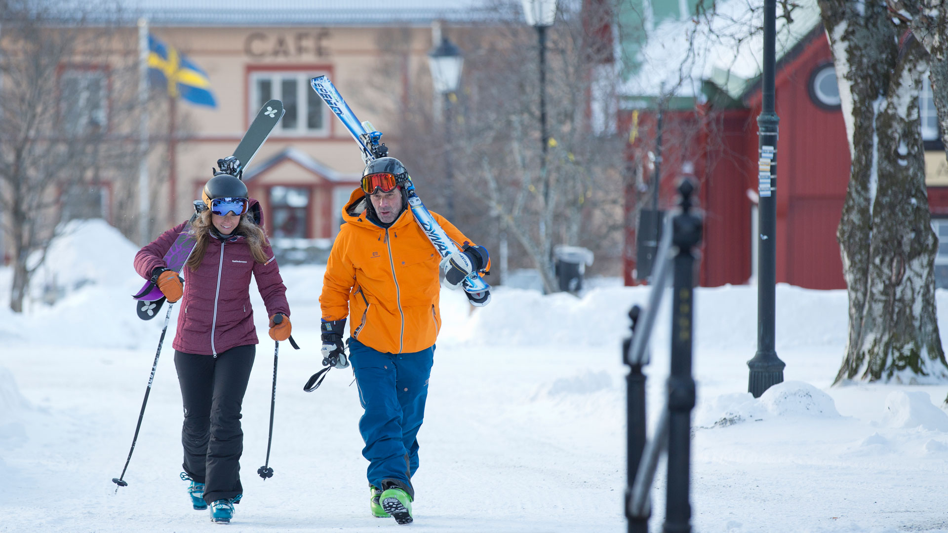 Två personer klädda i skidutrustning går genom en snöig by i Sverige. Svenska flaggan och ett café syns i bakgrunden