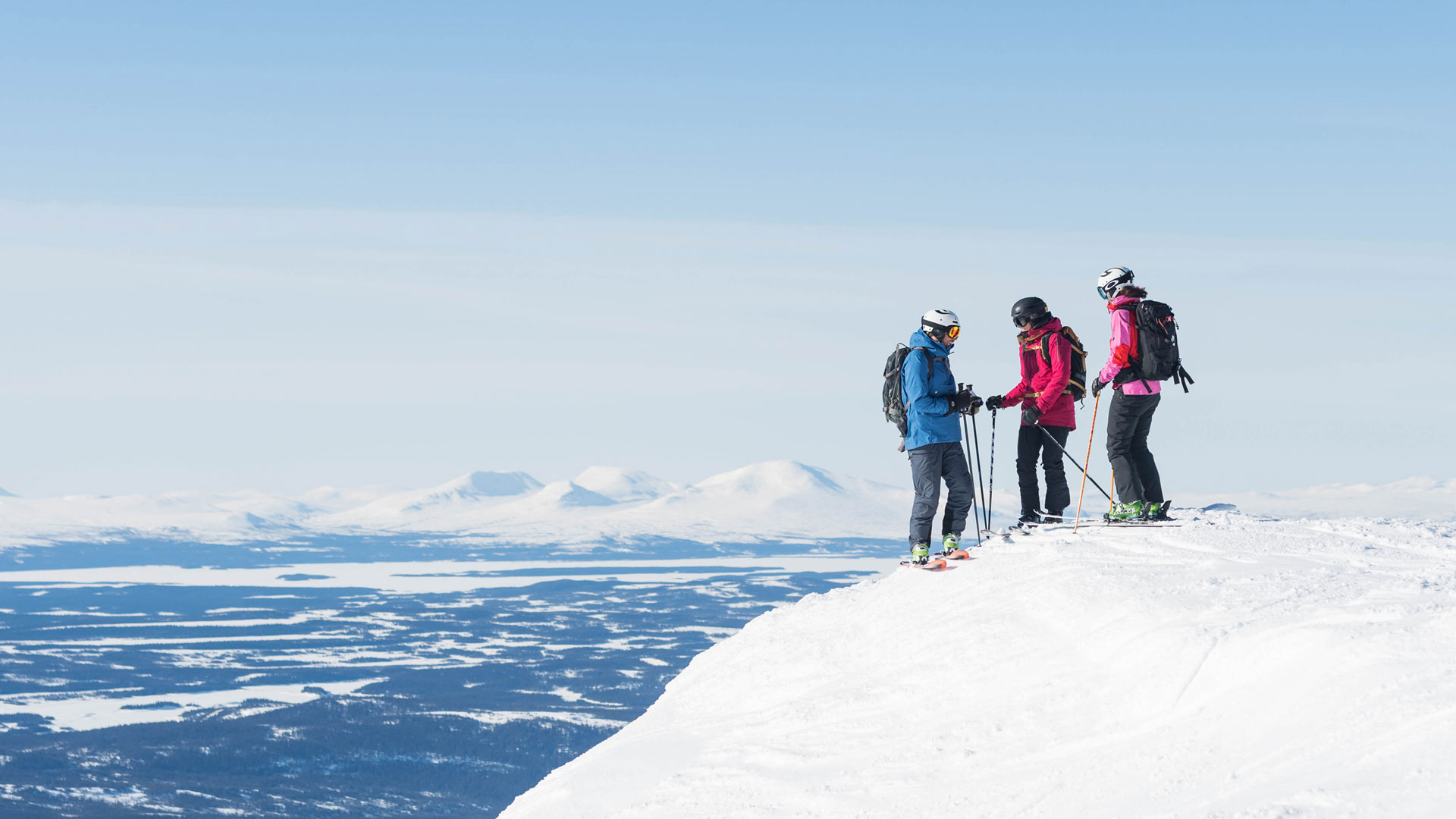 Tre skidåkare på snötäckt bergstopp i fjällandskap, klarblå himmel i bakgrunden