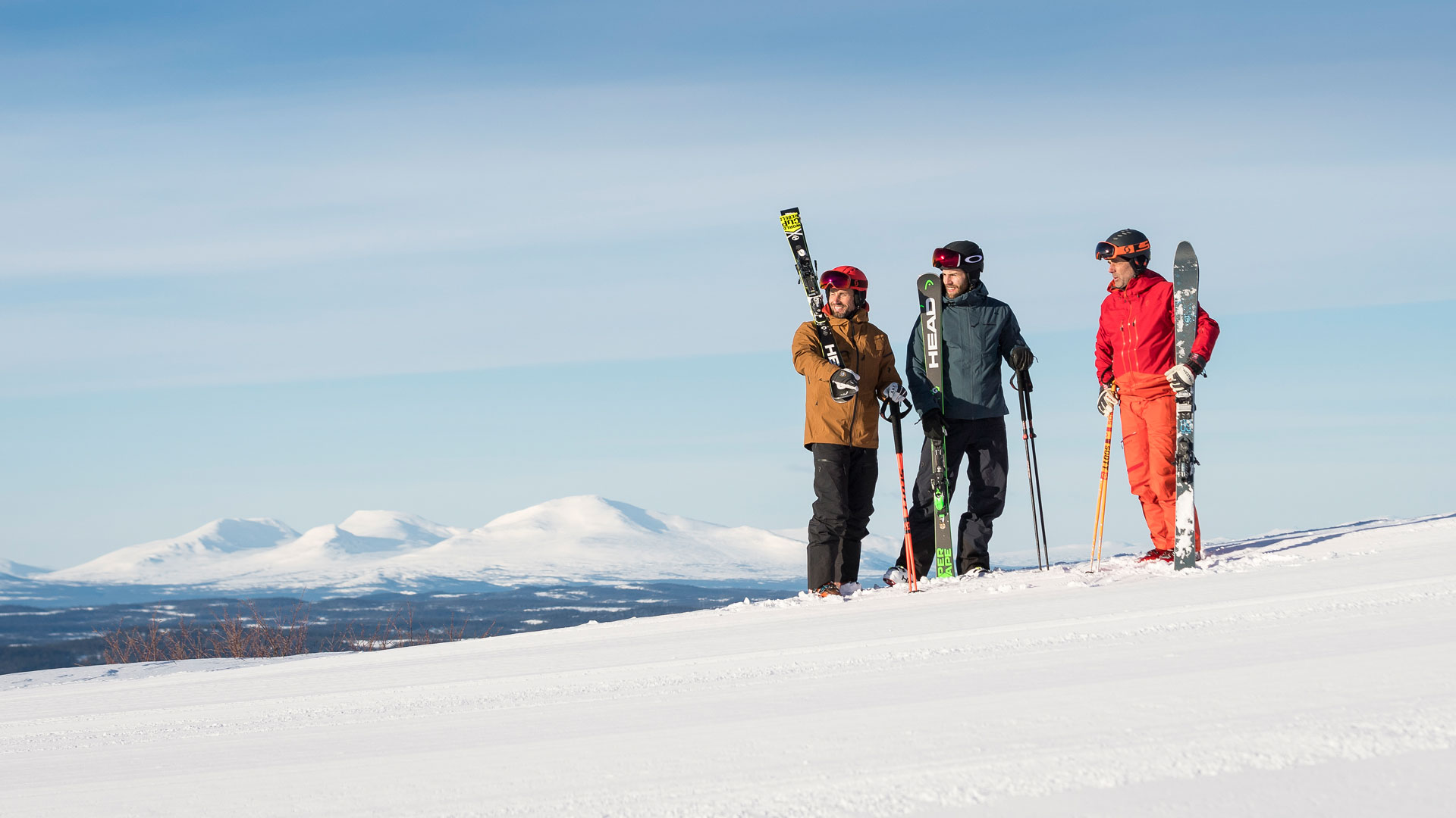Tre personer klædt i skitøj, der står med ski i hånden på en snedækket bjergskråning med snedækkede bjerge i baggrunden under en klar blå himmel