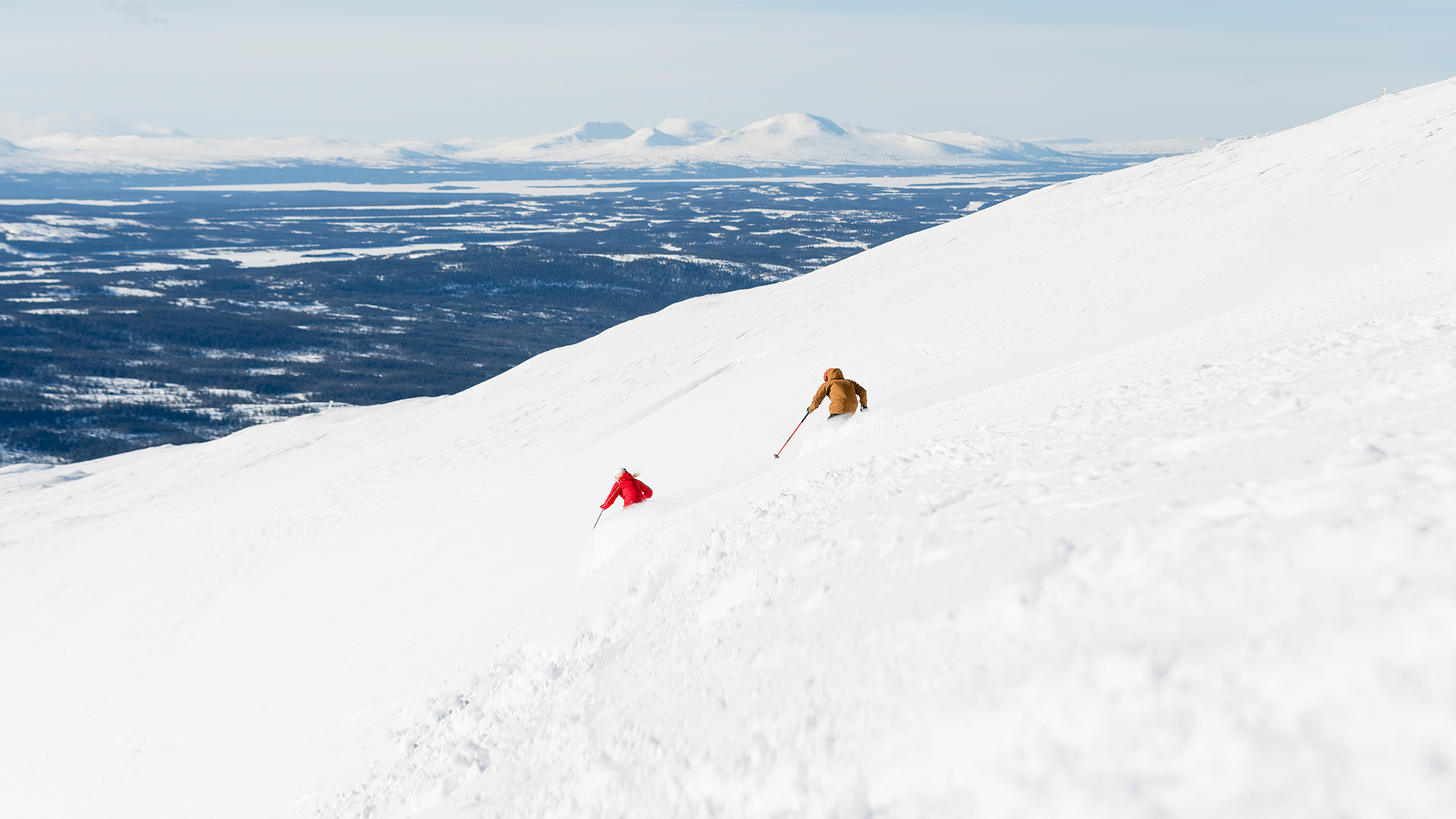 Två personer åker skidor nedför en snötäckt fjällsida i Sverige, med en vidsträckt utsikt över snölandskap och avlägsna berg i horisonten