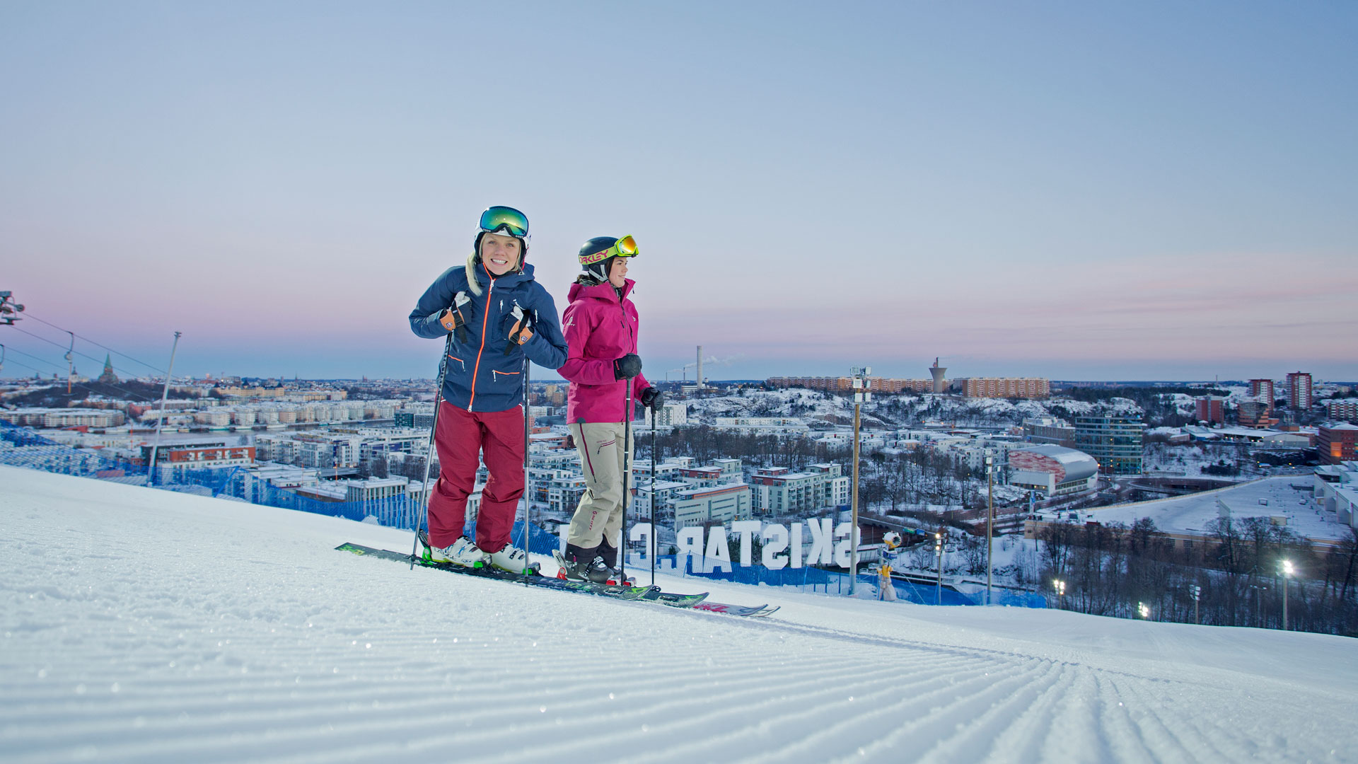 Två personer på skidor blickar ner från en skidbacke i Stockholm, med stadens silhuett i bakgrunden under en klar himmel