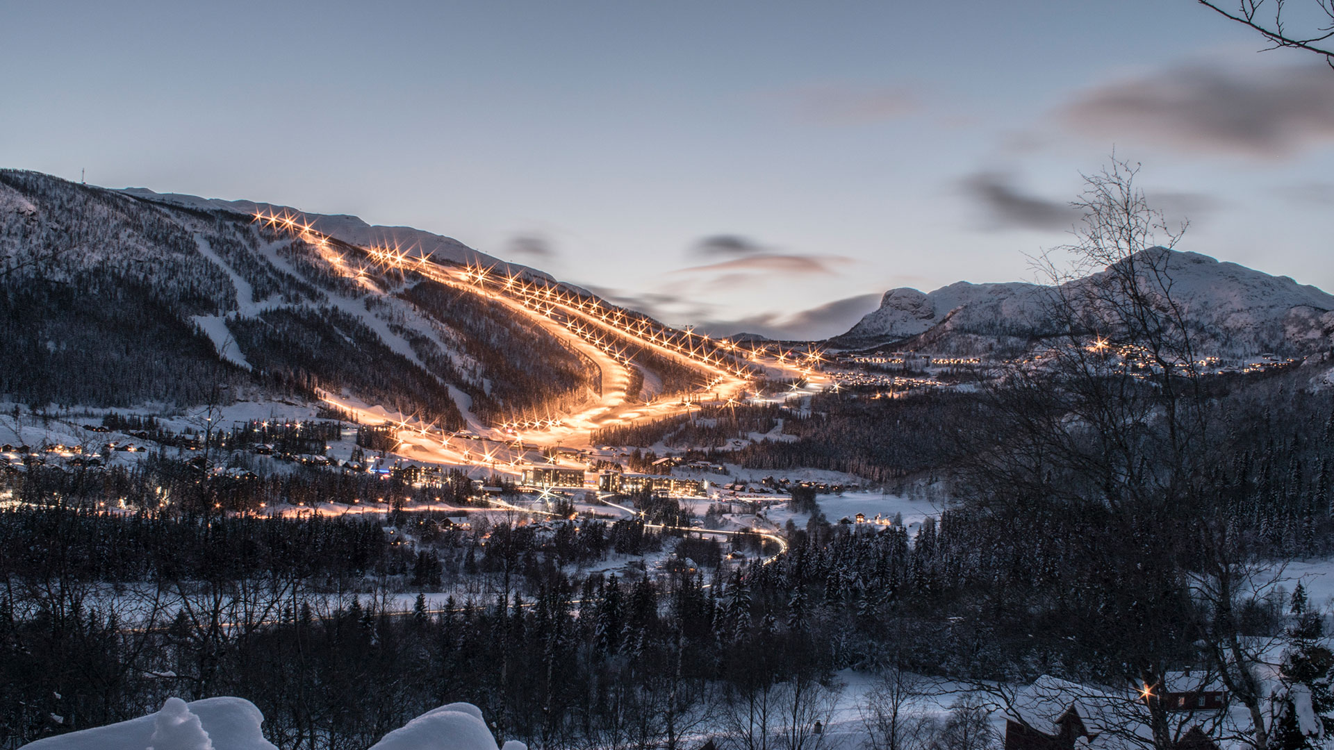 Kvällsbild av belyst skidbacke i snötäckt bergslandskap, omgiven av skog och små stugor under vintern