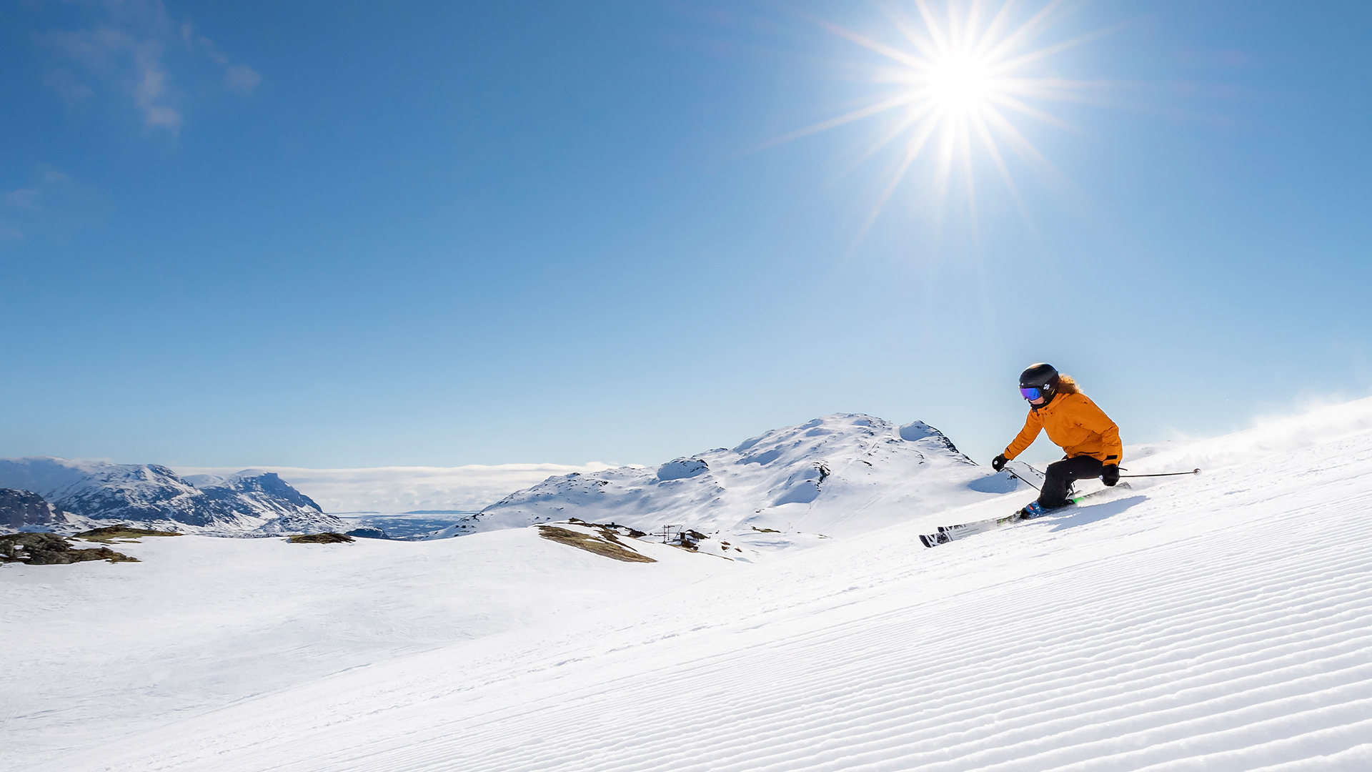 Person som åker skidor nedför en solig bergssluttning med klarblå himmel och snötäckta berg i bakgrunden