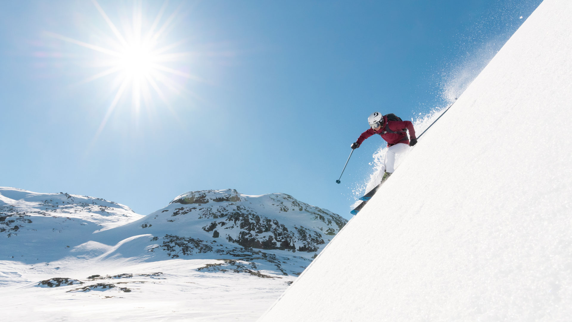 A skier in a red jacket is skiing down a snow-covered mountainside under a clear blue sky with the sun shining