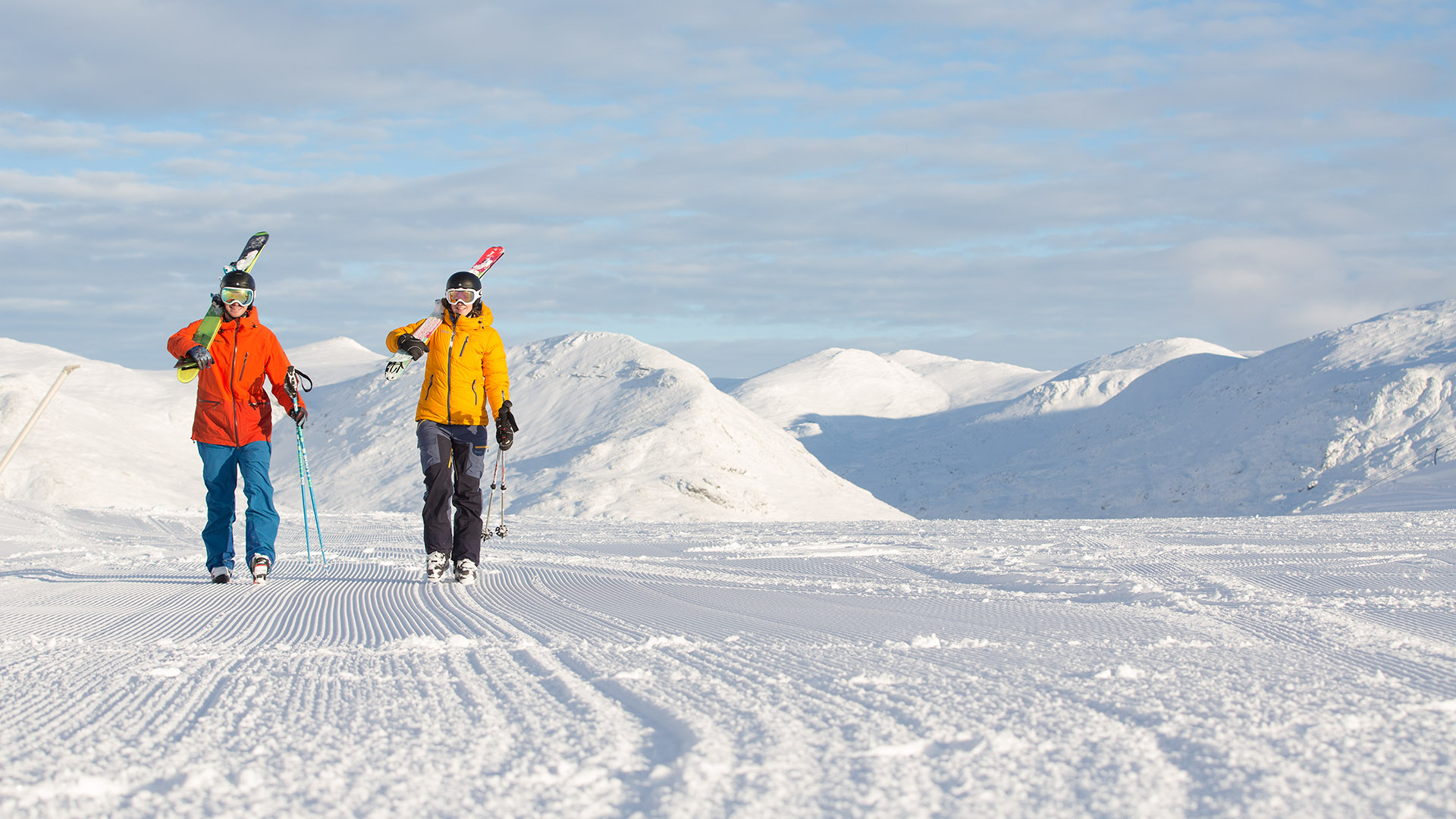 Två personer klädda i färgglada skiddräkter bär skidor på axlarna och går på en snötäckt backe med berg i bakgrunden under en klar himmel