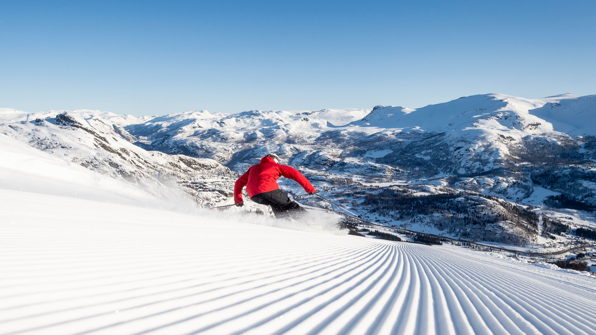 Person i röd jacka åker skidor nerför en preparerad skidbacke i ett snötäckt bergslandskap