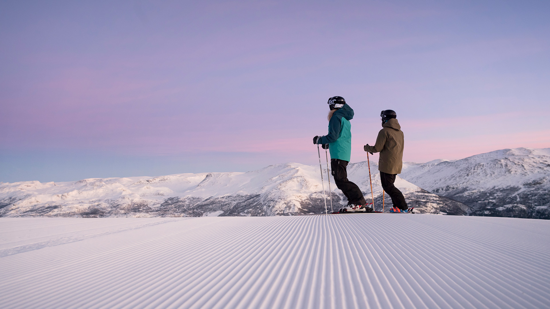 Two skiers on a groomed slope in snow-covered mountains, magical sunset in the background, Norway