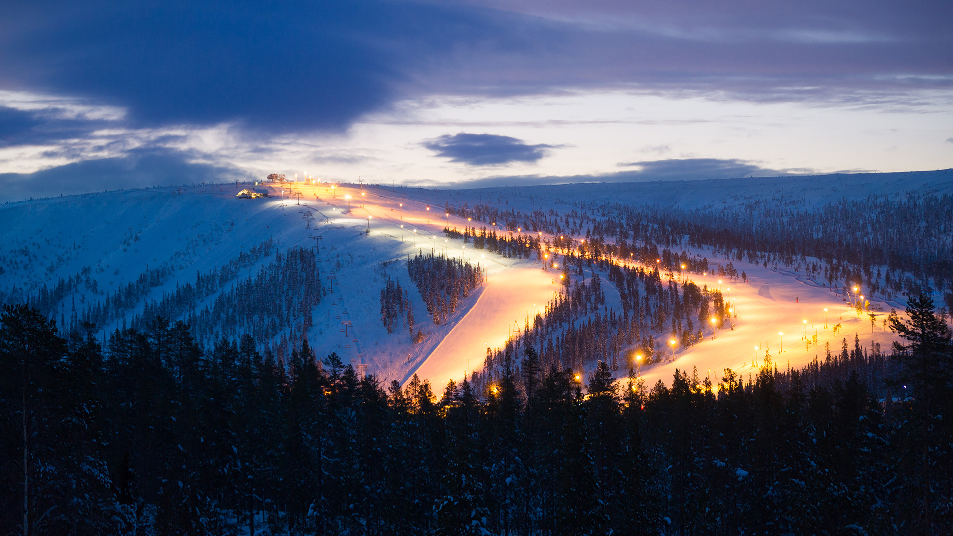 Nighttime view of a snow-covered ski slope illuminated with lights, surrounded by dense forest, under a cloudy sky