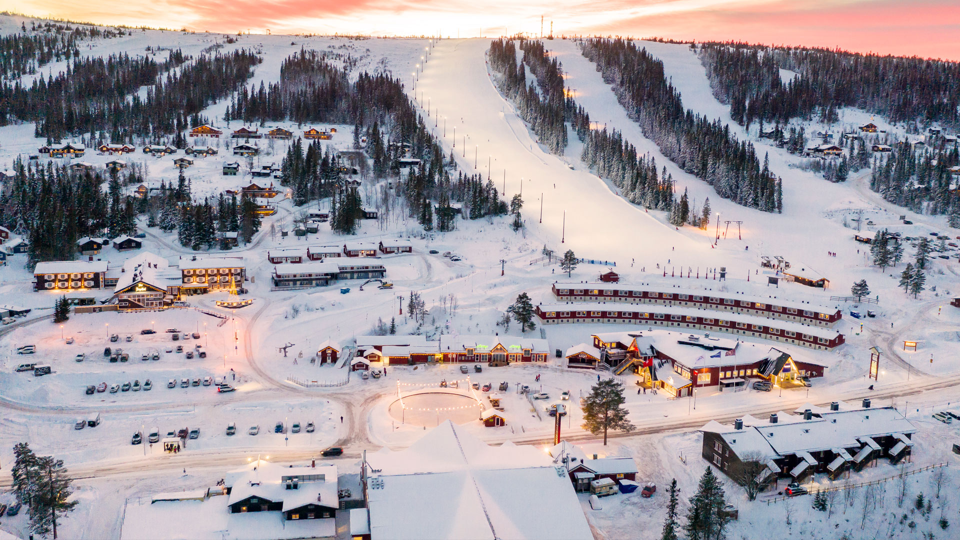 Landscape view of a winter resort with ski slopes, cabins, and cars under a colorful sunset, surrounded by snow-covered trees and mountains