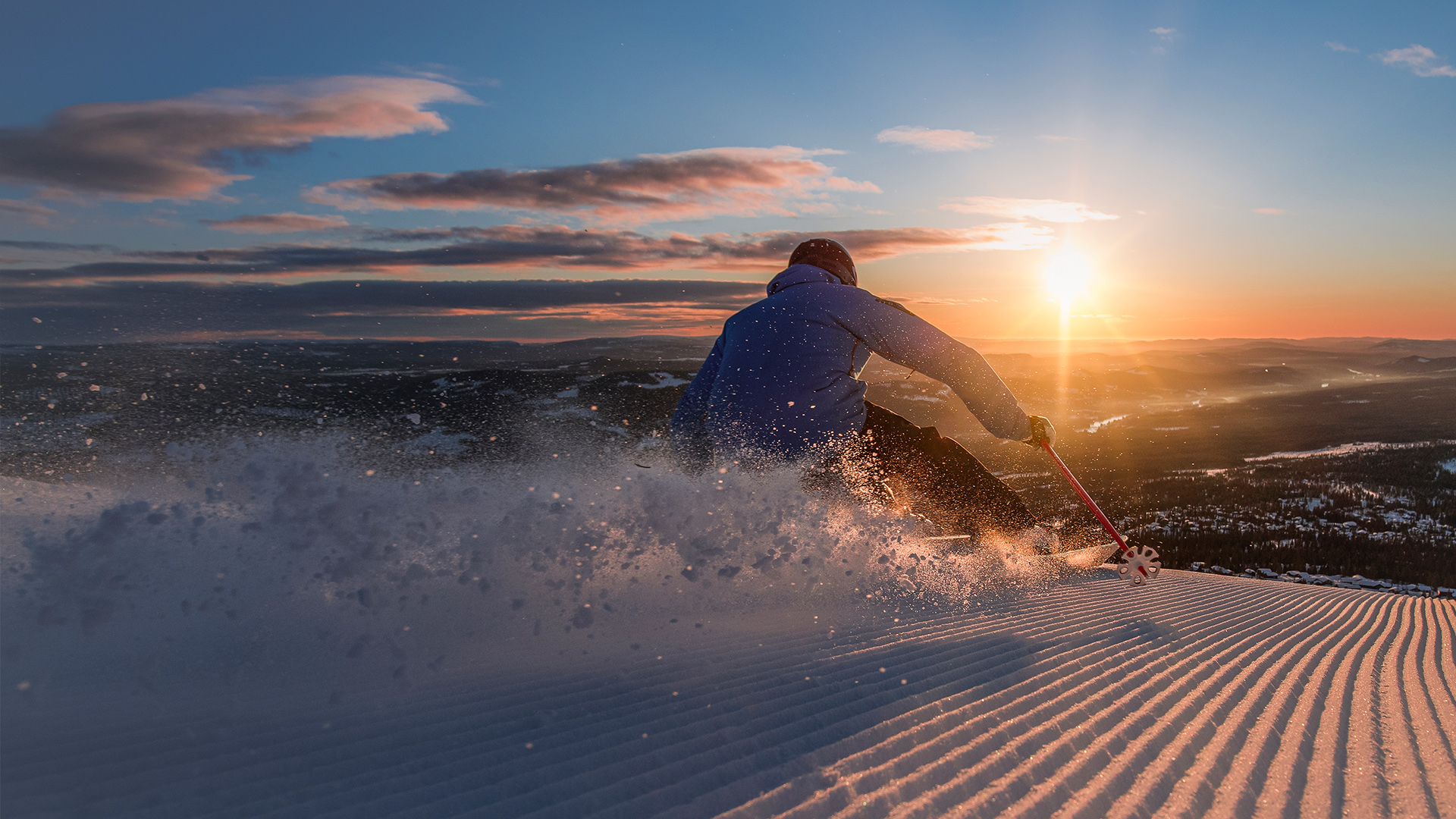Person åker skidor nerför en backe i solnedgången, med snö som virvlar i luften och ett vackert fjällandskap i bakgrunden