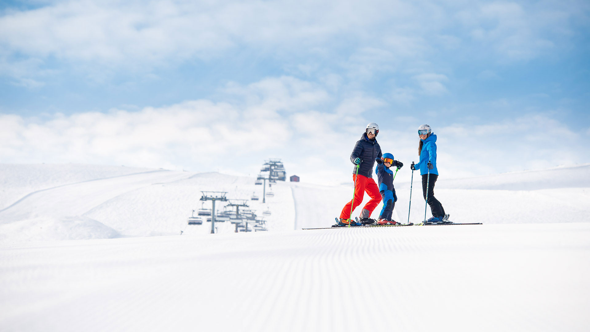 A family is skiing in a sunny winter landscape with snow-covered mountains and ski lifts in the background