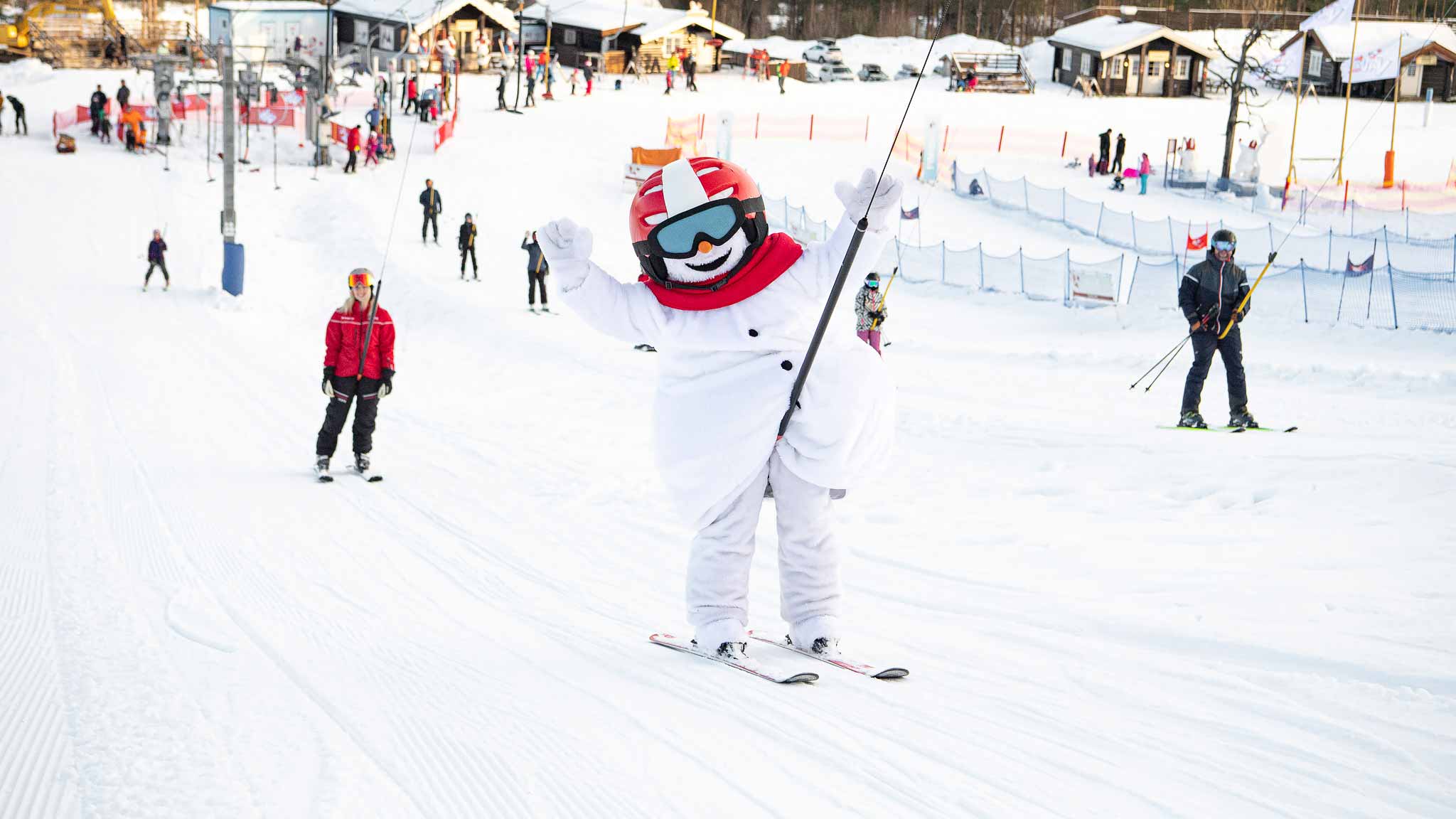 Valle (snögubbe-maskot) åker skidor i en livlig skidort med andra skidåkare i bakgrunden, omgiven av snötäckta stugor och liftar