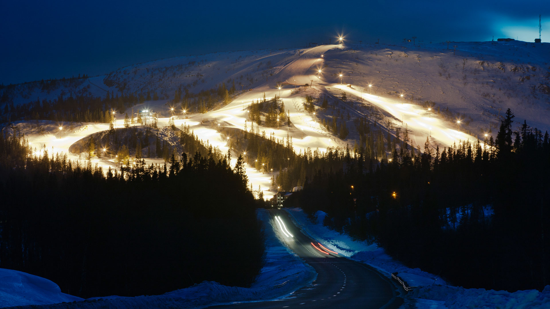A night-lit ski slope in a snowy landscape with pine forest in the foreground and a road at dusk, perfect for evening skiing in the mountains