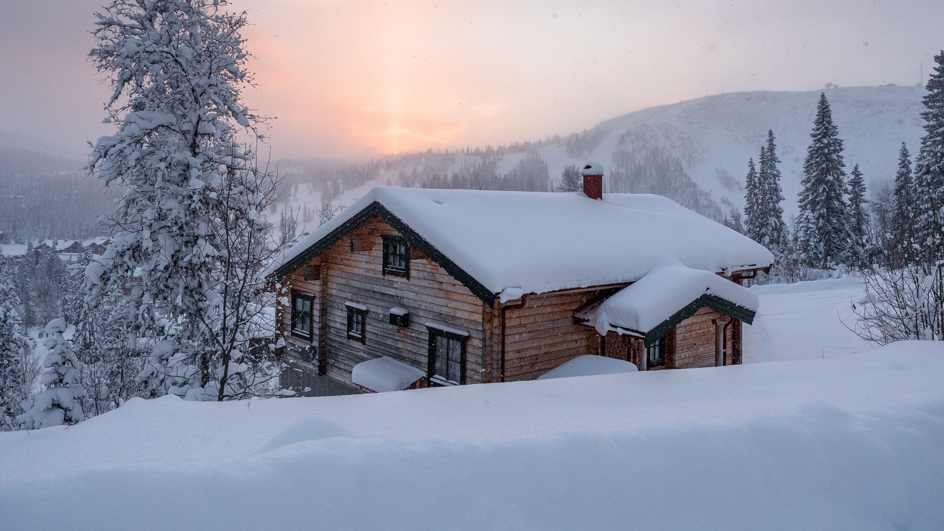 Trähus i ett snötäckt vinterlandskap med solnedgång i bakgrunden, omgiven av skog och berg