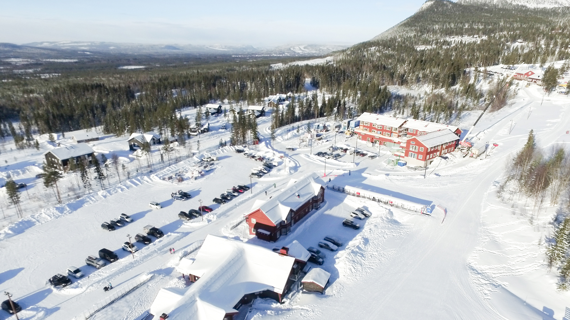 Flygfoto över en snötäckt skidanläggning i Björnrike, Sverige. Flera byggnader och parkerade bilar syns, med snötäckta berg i bakgrunden