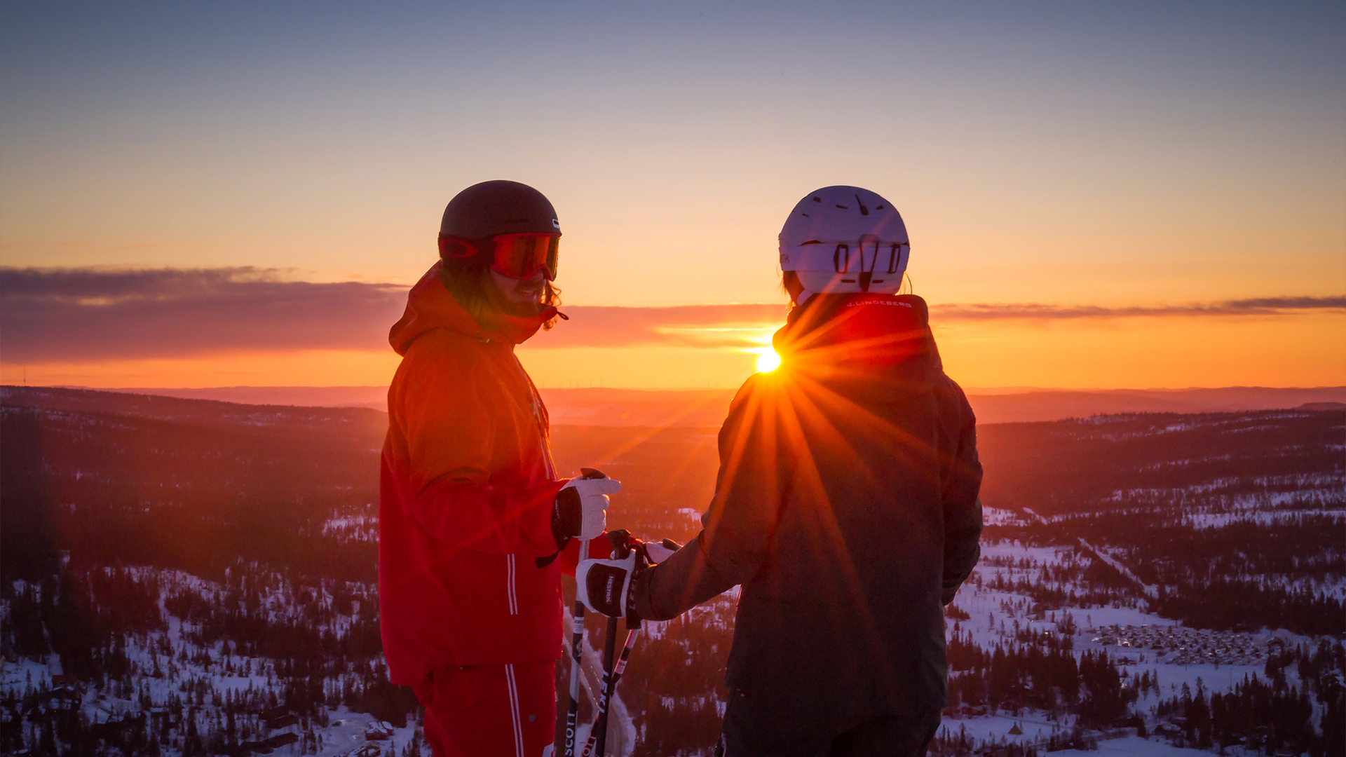 Two skiers at sunset on a snow-covered mountain, with sunbeams shining through their silhouettes