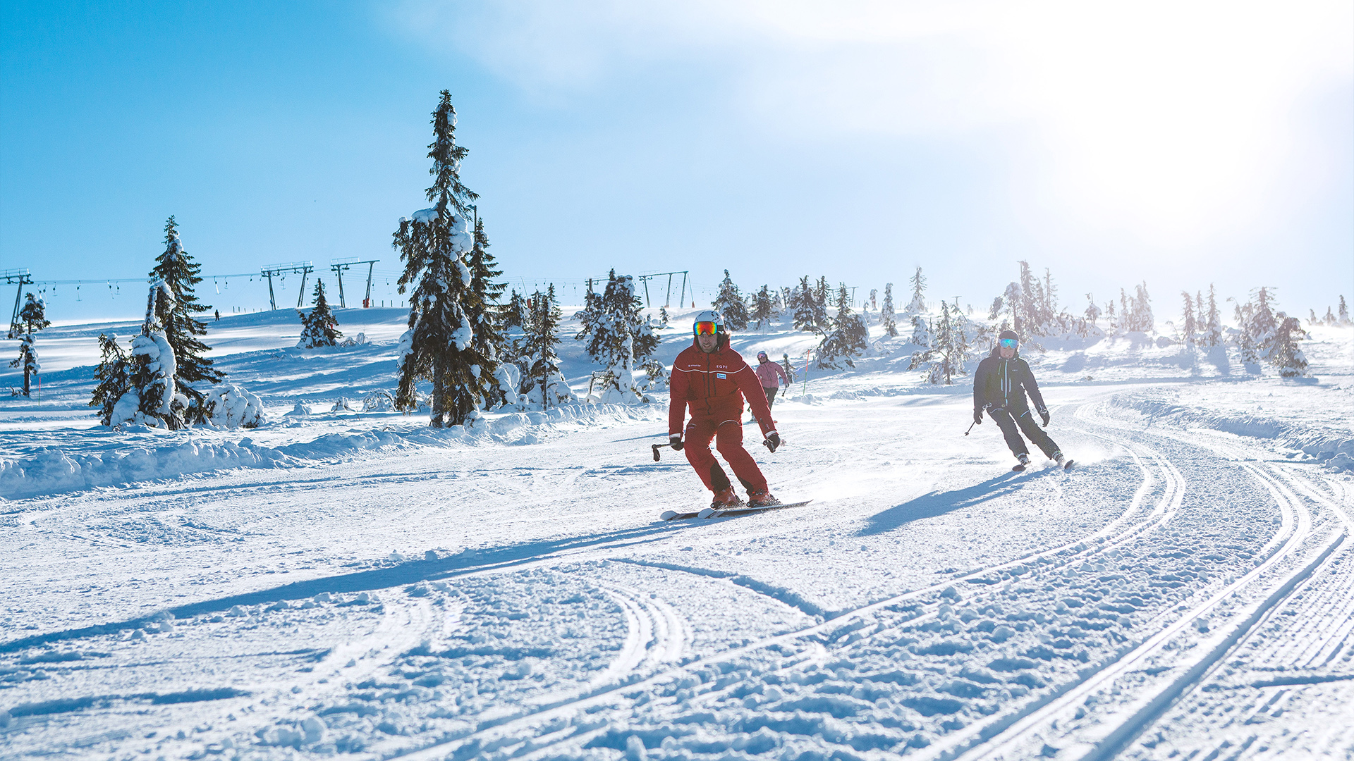 Skidlärare i röda kläder åker med elev i svarta kläder nedför en solig fjällsluttning, omgivna av snöklädda träd och liftar i bakgrunden