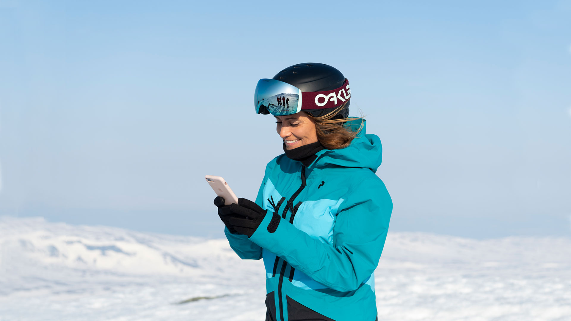 Person klädd i blå skidjacka och hjälm med googles, står i ett snölandskap och använder en smartphone. Solig vinterdag