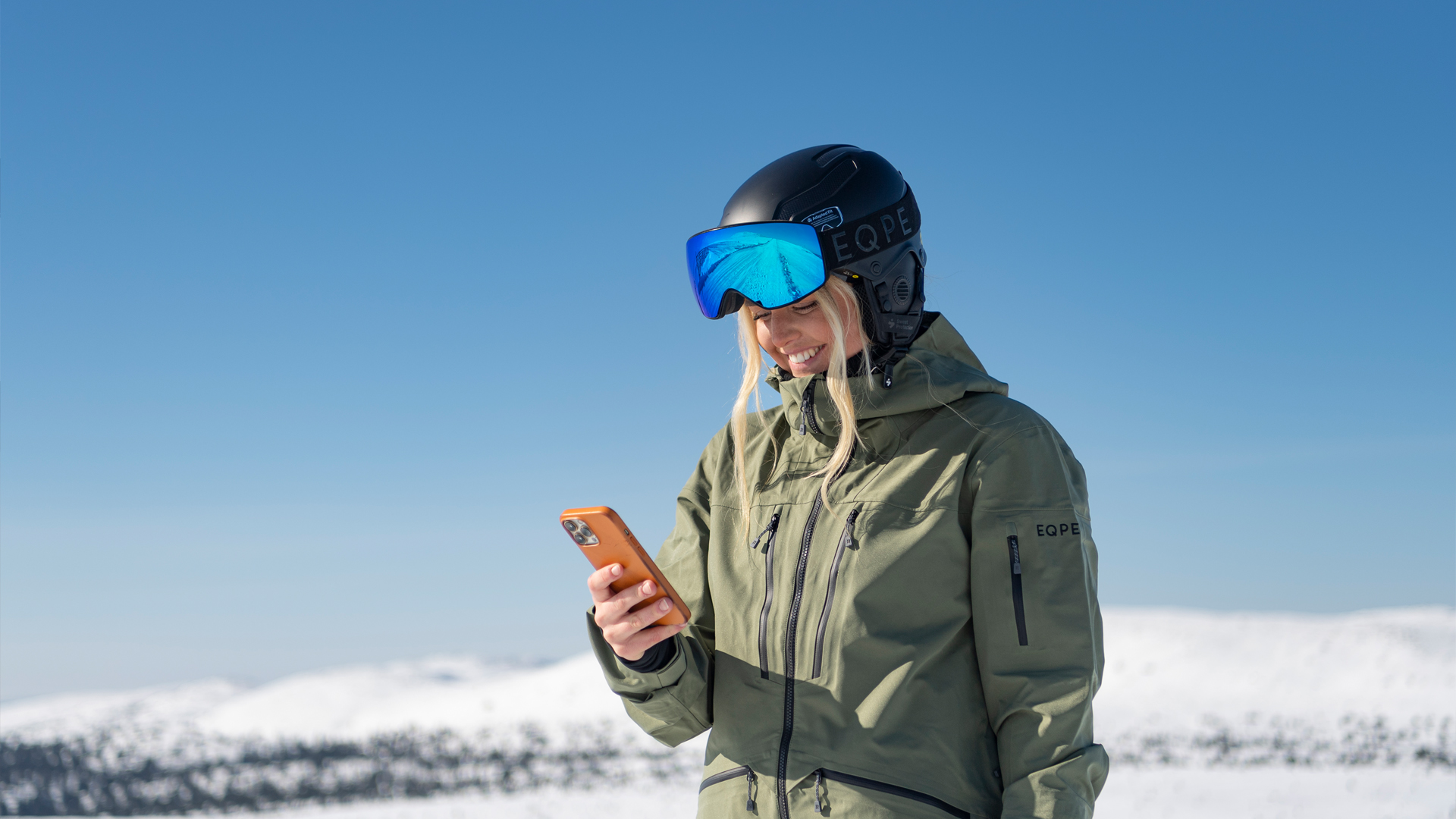 Kvinna i skidkläder med hjälm och skidglasögon använder mobiltelefon på snötäckt berg med klarblå himmel