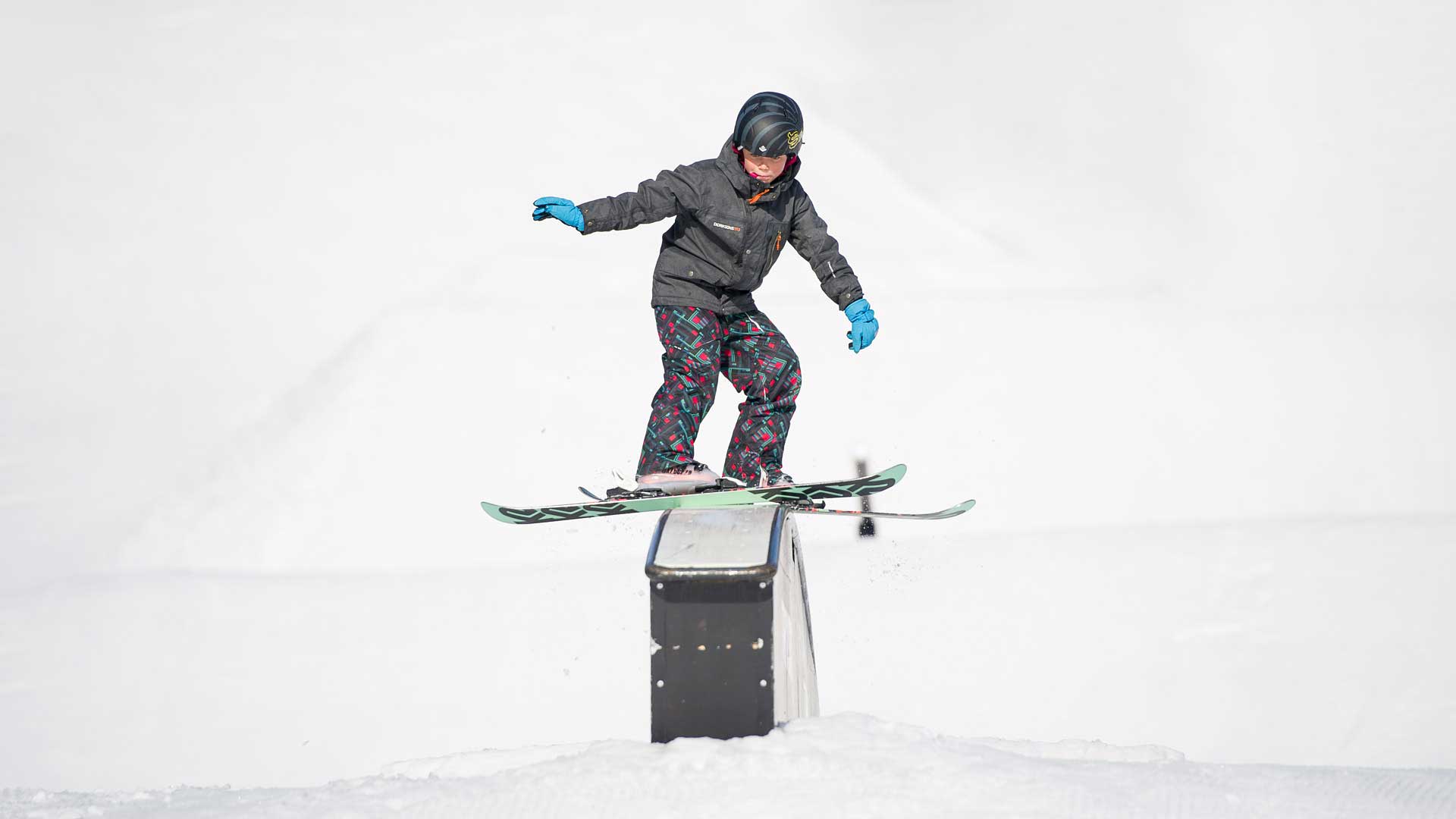En person står på ski på en rail i en snødekt terrengpark, iført hjelm og fargerike skidresser