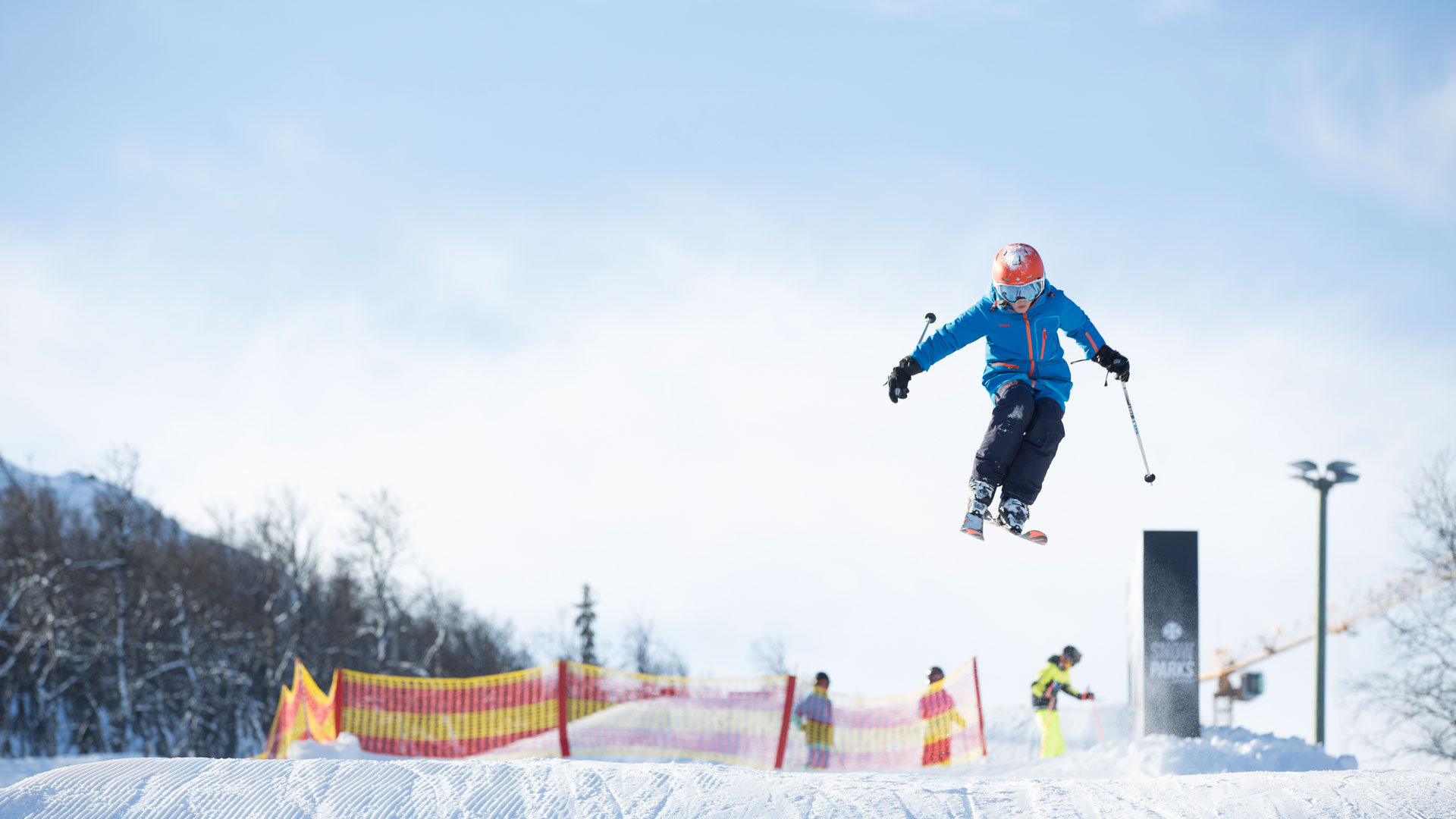 Person som utfører et høyt hopp på ski i et vinterlandskap, med solskinn og snødekte bakker i bakgrunnen