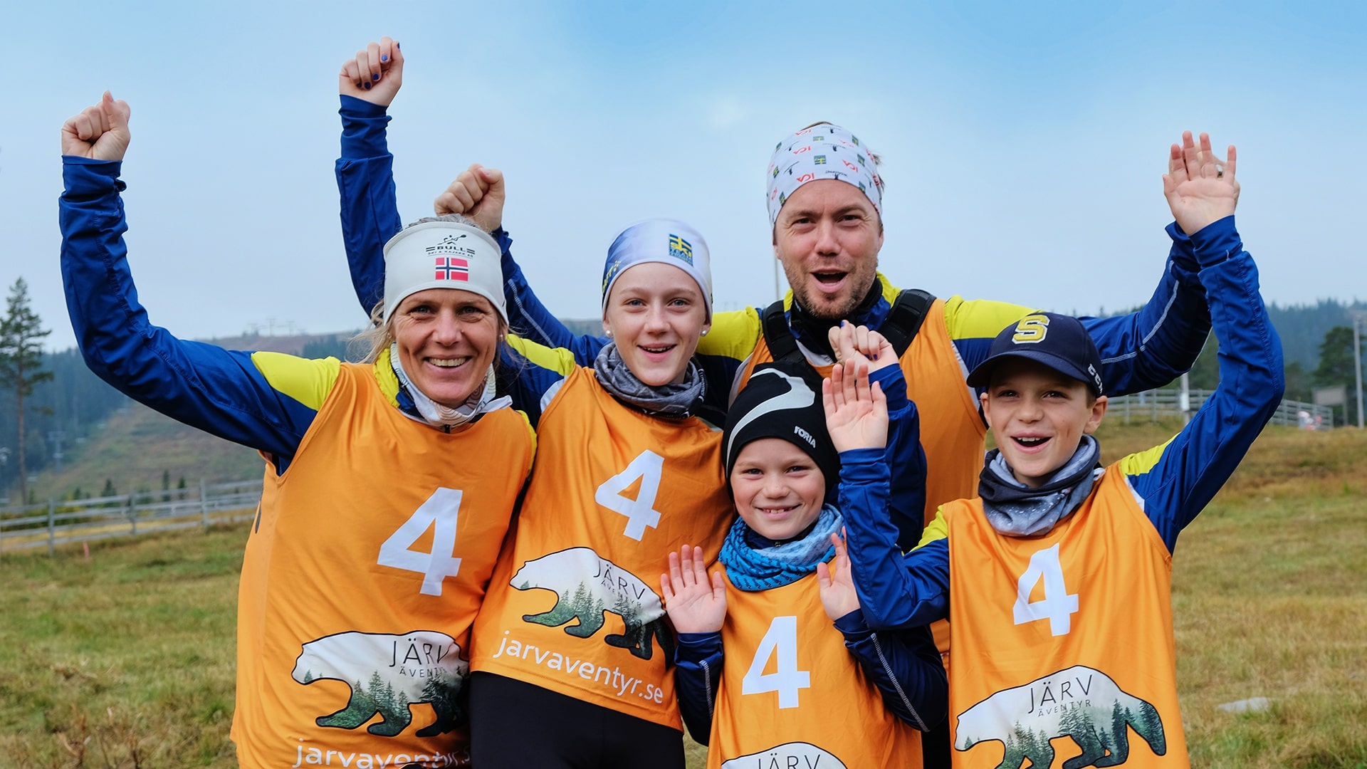 Glada deltagare med nummerlappar på Järväventyr, klädda i orange västar och mössor, poserar med upplyfta händer i skogsmiljö