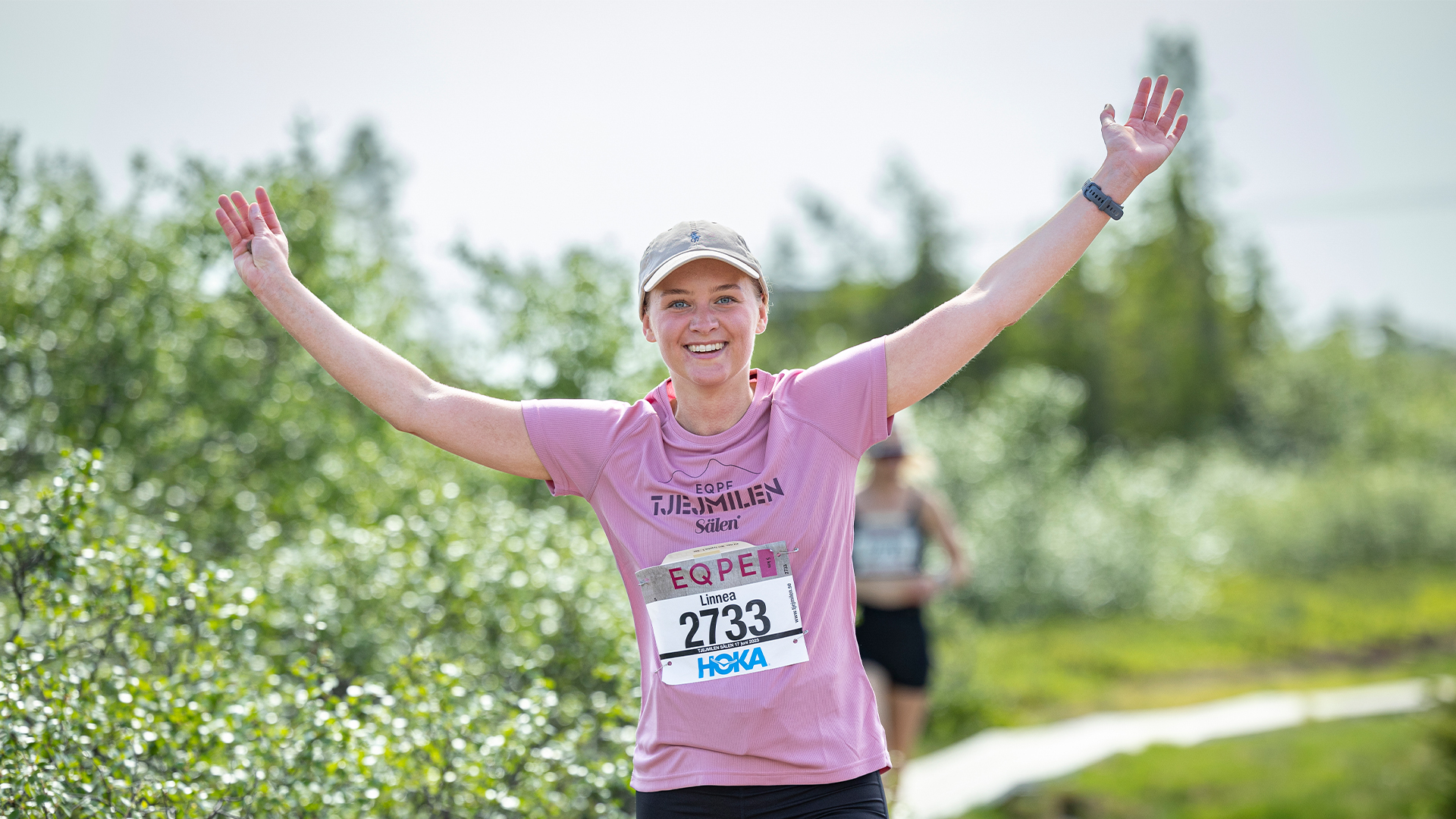 Person som springer Tjejmilen med armarna höjda, glad uttryck, klädd i rosa tröja och keps, grönskande bakgrund