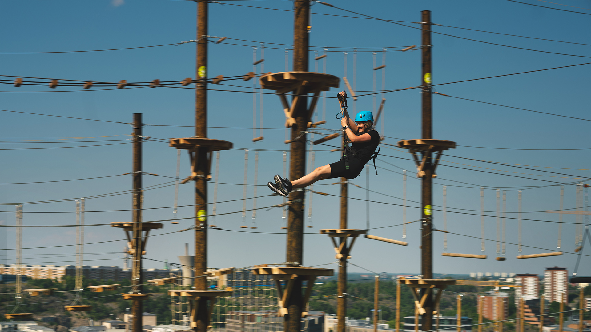 Person i hjälm åker zipline i en klätterpark utomhus med stadsmiljö i bakgrunden