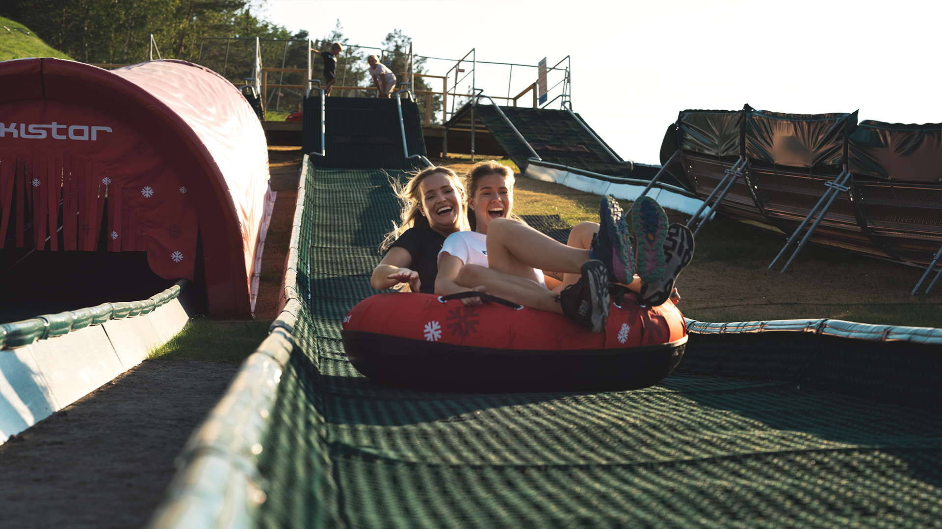 Två personer åker Mountain Tube i sommarpark, skrattar och har roligt i en uppblåsbar ring på en bana med grön matta
