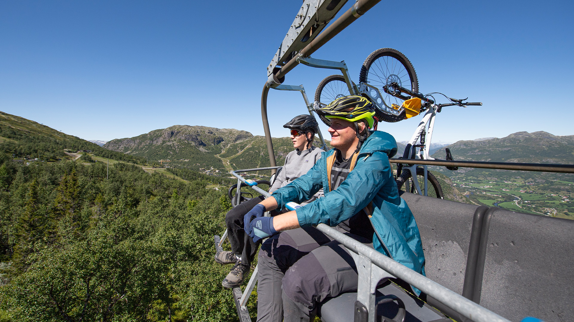 Två personer i cykelgläder och hjälmar åker en stollift med mountainbikes i bakgrunden, omgivna av grönskande berg och klarblå himmel