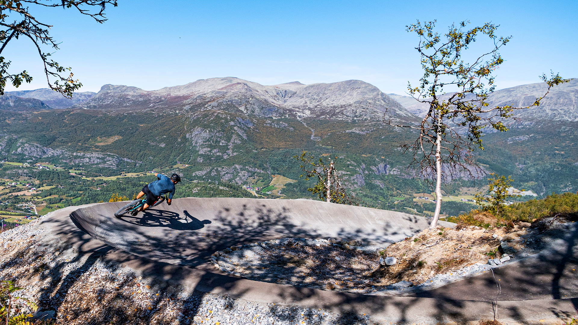 Cyklist åker nedför bergsstig med storslagen utsikt över fjäll i bakgrunden, solig dag