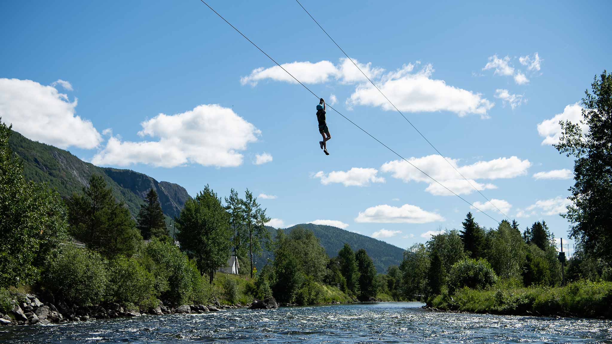 Person som åker zipline över flod med naturskön utsikt av skog och berg i bakgrunden under en solig dag