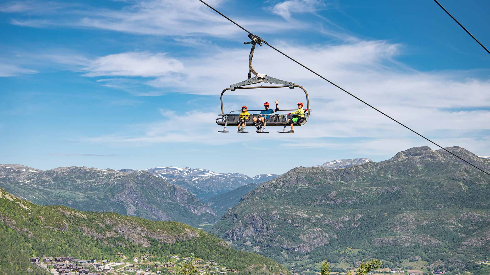 Tre personer i en sittlift ovanför majestätiska berg i soligt väder, med panoramautsikt över dalen och snötäckta toppar i bakgrunden