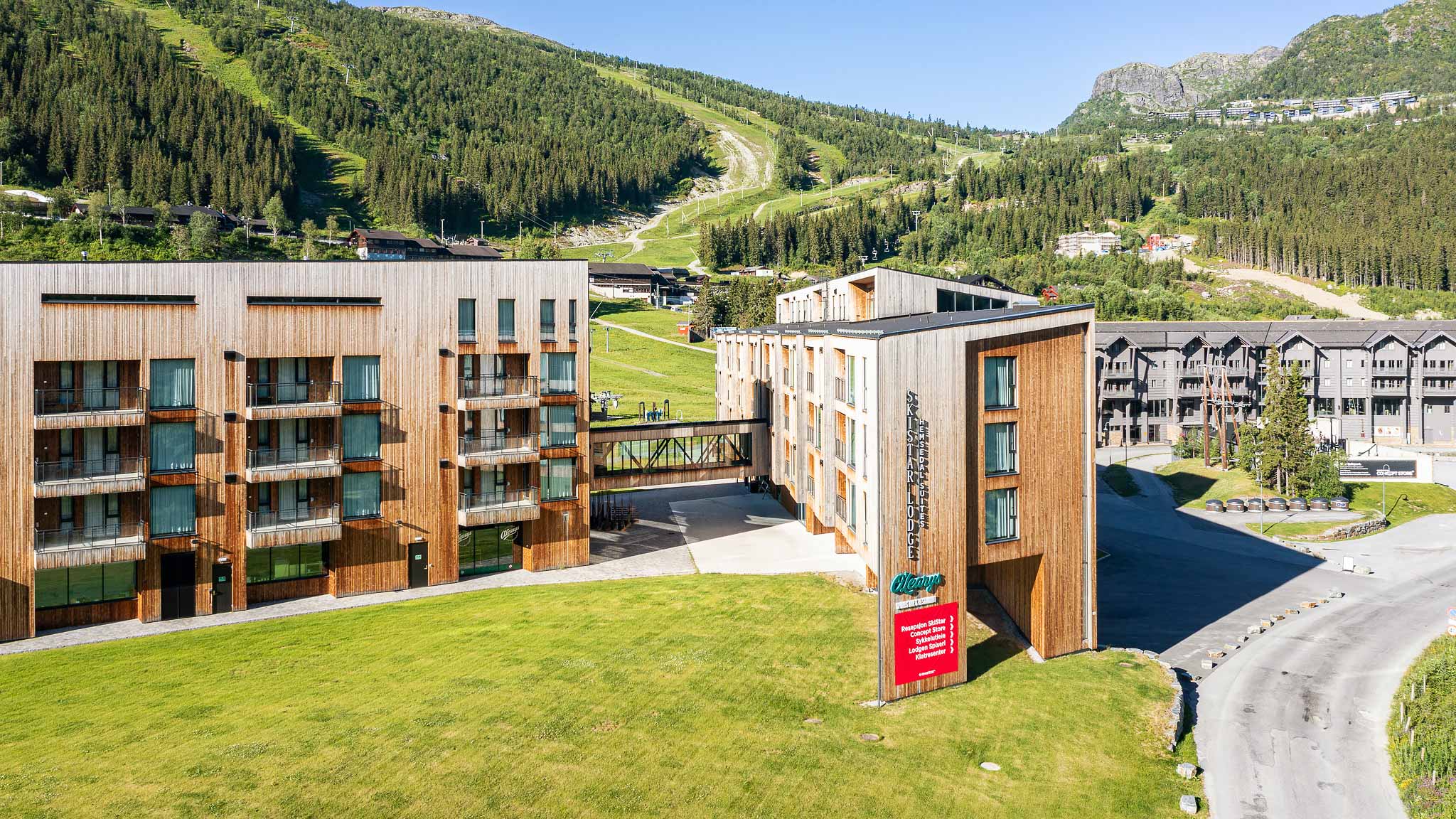 Hotellbyggnad med modern design, omgiven av grönskande bergslandskap och skidbackar i bakgrunden, under en klarblå himmel