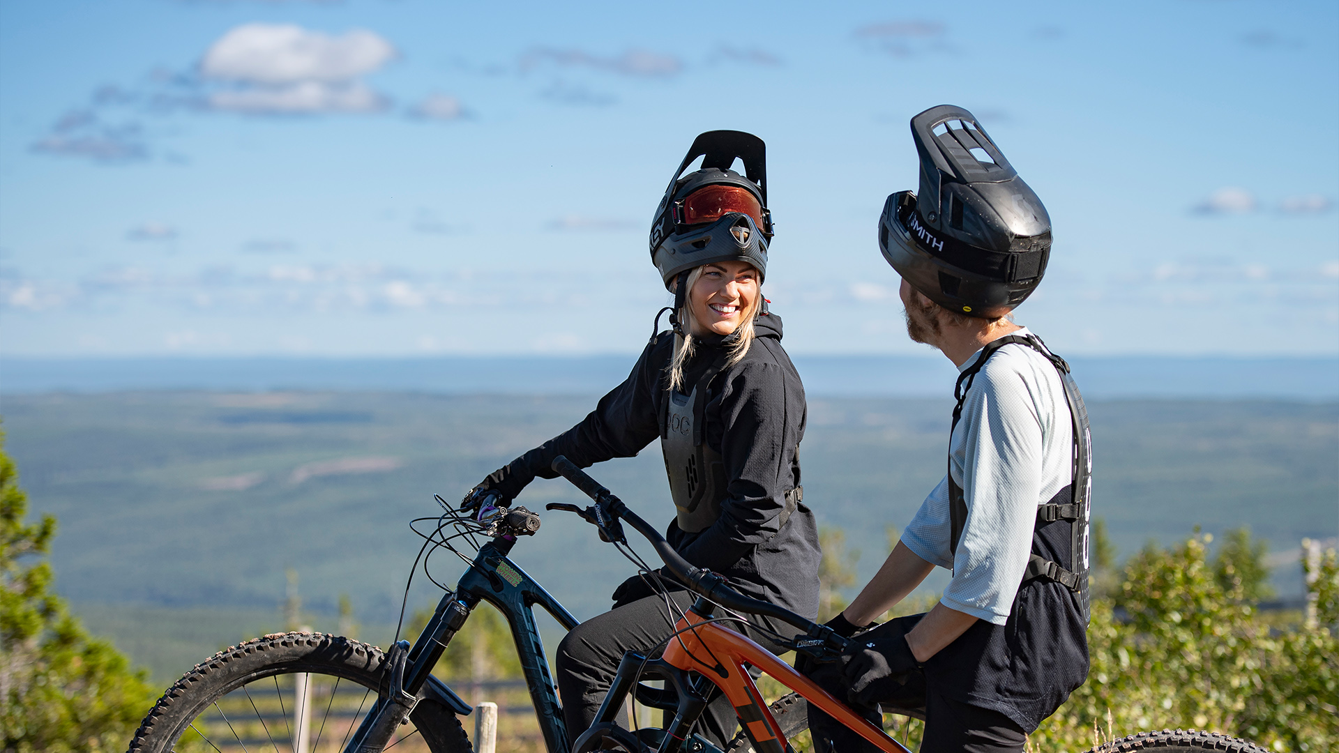 Två cyklister med hjälmar pratar och ler medan de sitter på sina mountainbikes, med en vidsträckt utsikt över ett landskap i bakgrunden