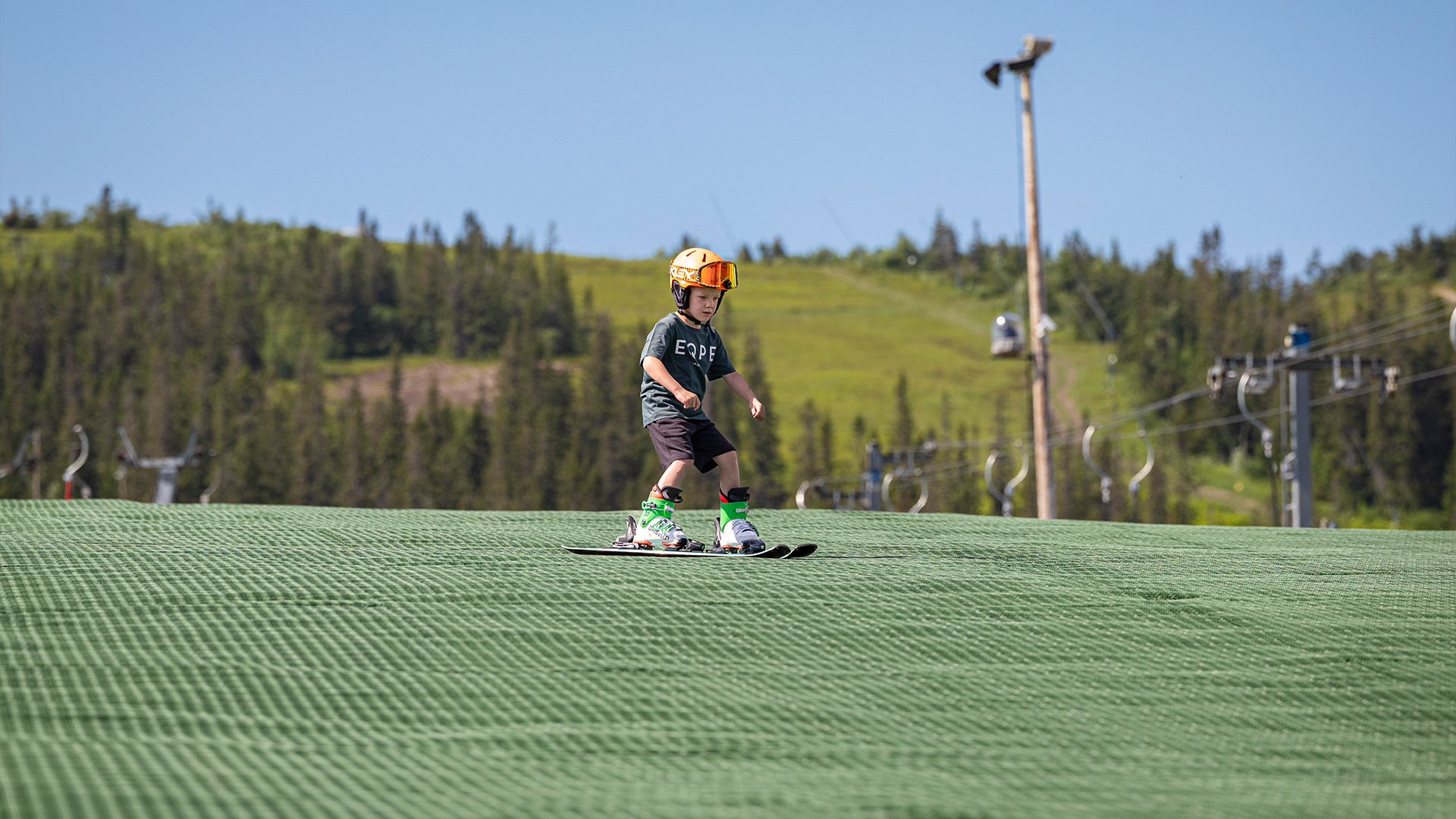 Ungt barn åker skidor på konstgjord skidmatta i somrigt landskap, iförd hjälm och skidutrustning. Skidliftar och grönskande skog i bakgrunden
