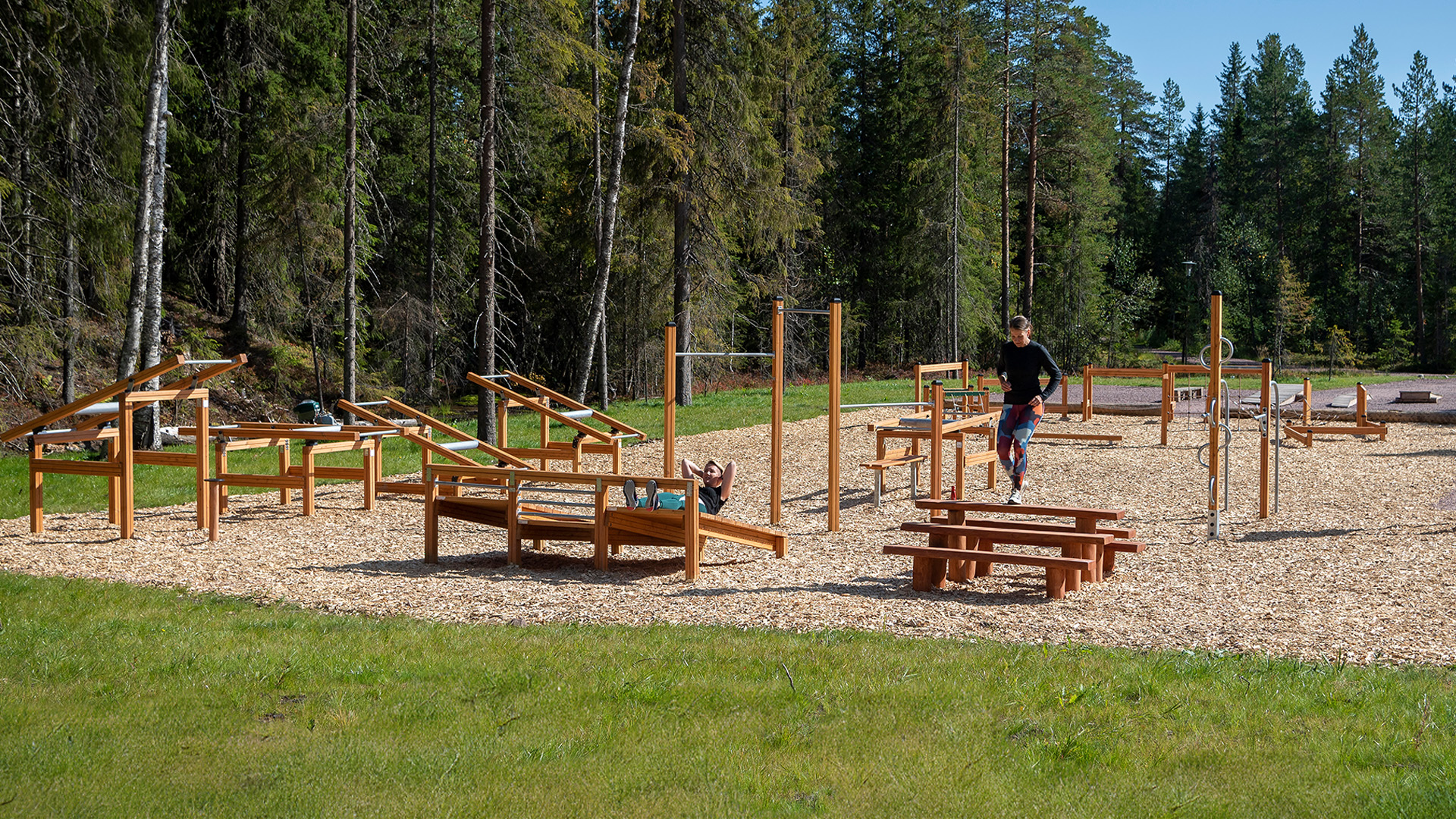 Utomhusgym i skogsmiljö med träningsutrustning i trä och en person som tränar, omgiven av grönska och träd i bakgrunden