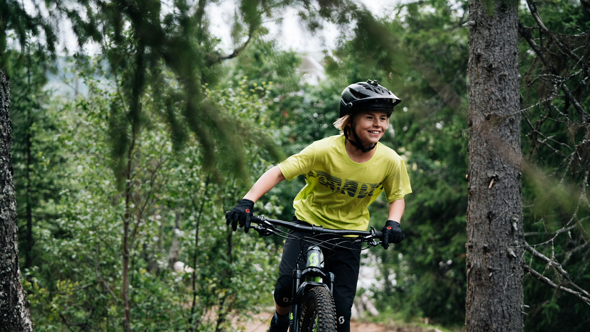 Ung cyklist med hjälm och gul tröja på mountainbike i skogen, omgiven av gröna träd
